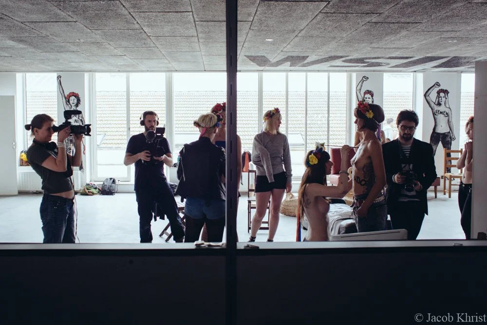 Un groupe de personnes en train de se faire photographier et filmer dans une pièce lumineuse avec de grandes fenêtres. Deux femmes sont nues et portent des couronnes de fleurs, une d'elles est en train de se faire maquiller ou ajuster. Quelques dessi
