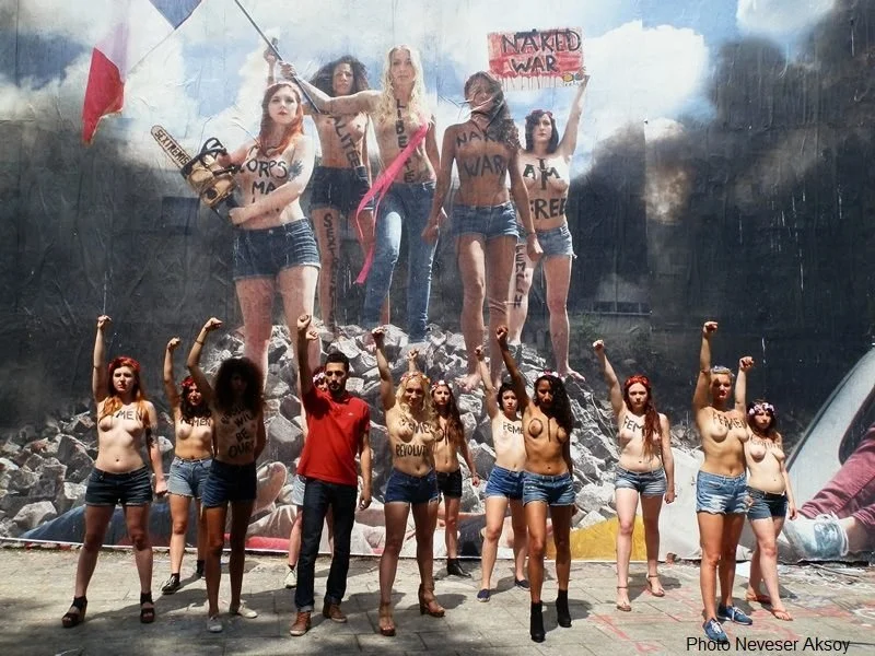 Groupe de personnes manifestant, dont certaines ont le corps peint avec des mots comme 'FEMINISTE' et 'REVOLU' et tiennent des protestations contre la guerre et la domination masculine, devant une affiche géante en arrière-plan.