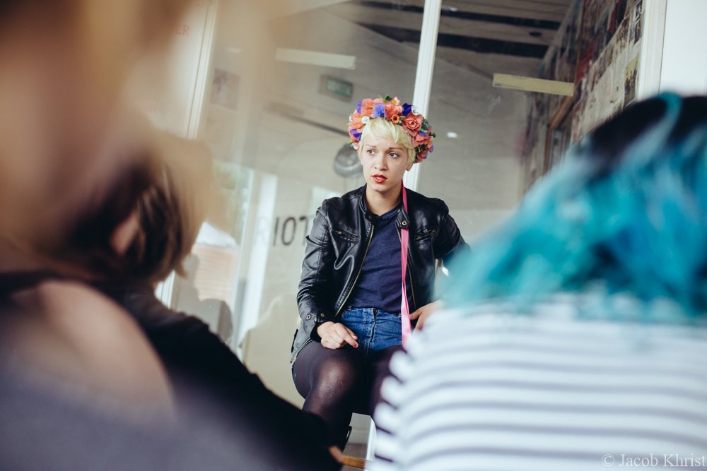 Une jeune femme avec un manteau en cuir, coiffée d'une couronne de fleurs, assise et parlant avec deux autres personnes dans un espace intérieur.