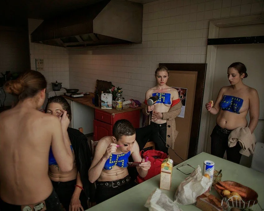 Groupe de jeunes femmes et un jeune homme dans une cuisine, certaines portant des vestes de l'Union européenne ou de l'Europe, d'autres non, en train de se préparer ou de manger.