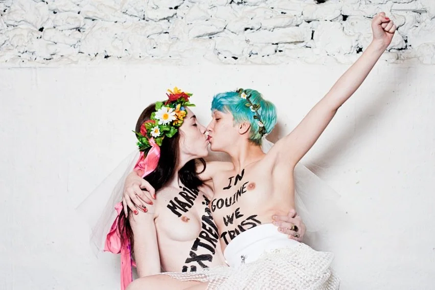 Deux femmes nues portant des couronnes de fleurs, s'embrassant et se tenant dans un mur blanc.