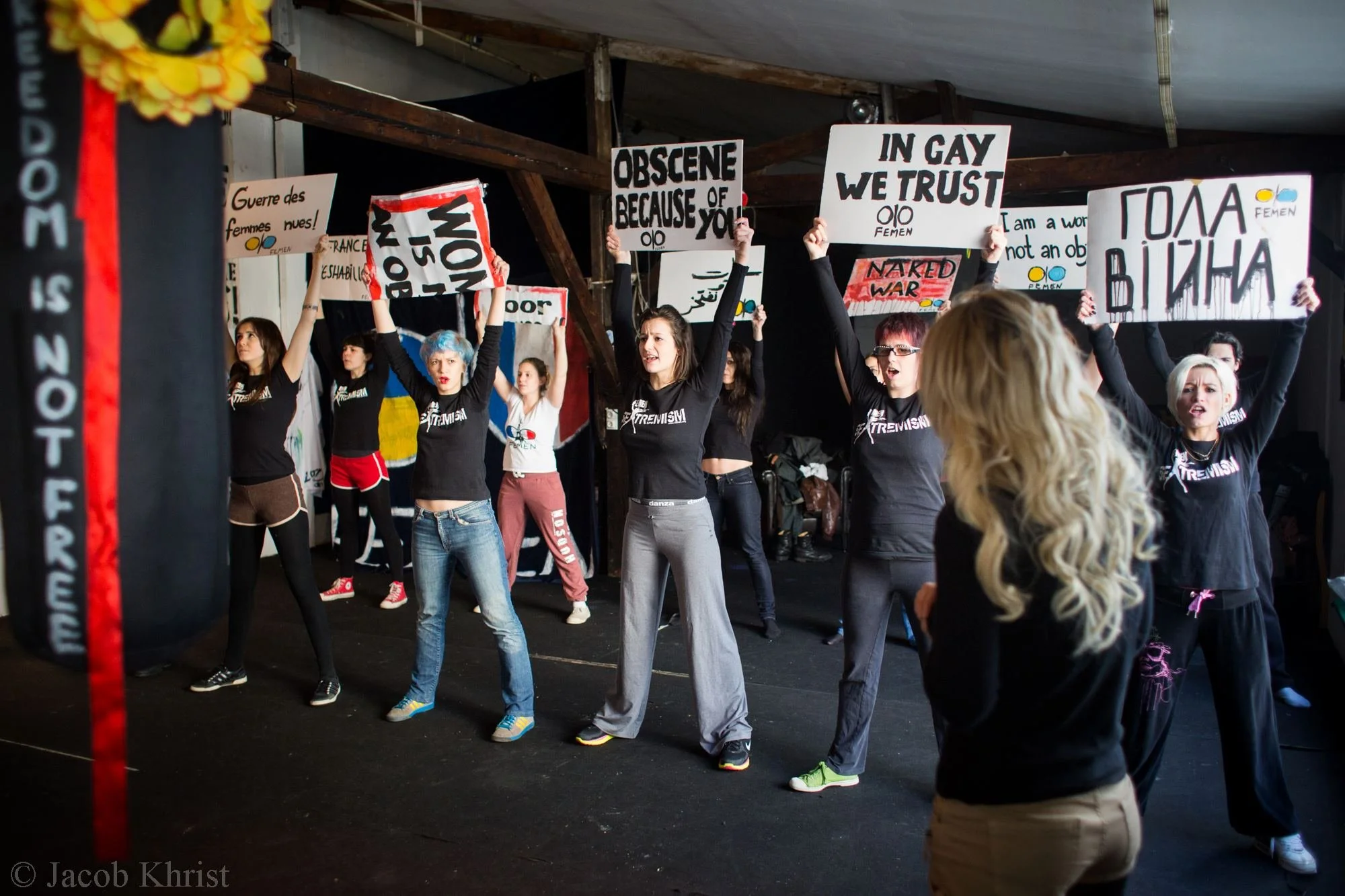 Jeunes femmes manifestant avec des pancartes en français et en anglais lors d'une protestation pour les droits des femmes et contre l'activisme extrémiste, dans une salle avec des poutres en bois.