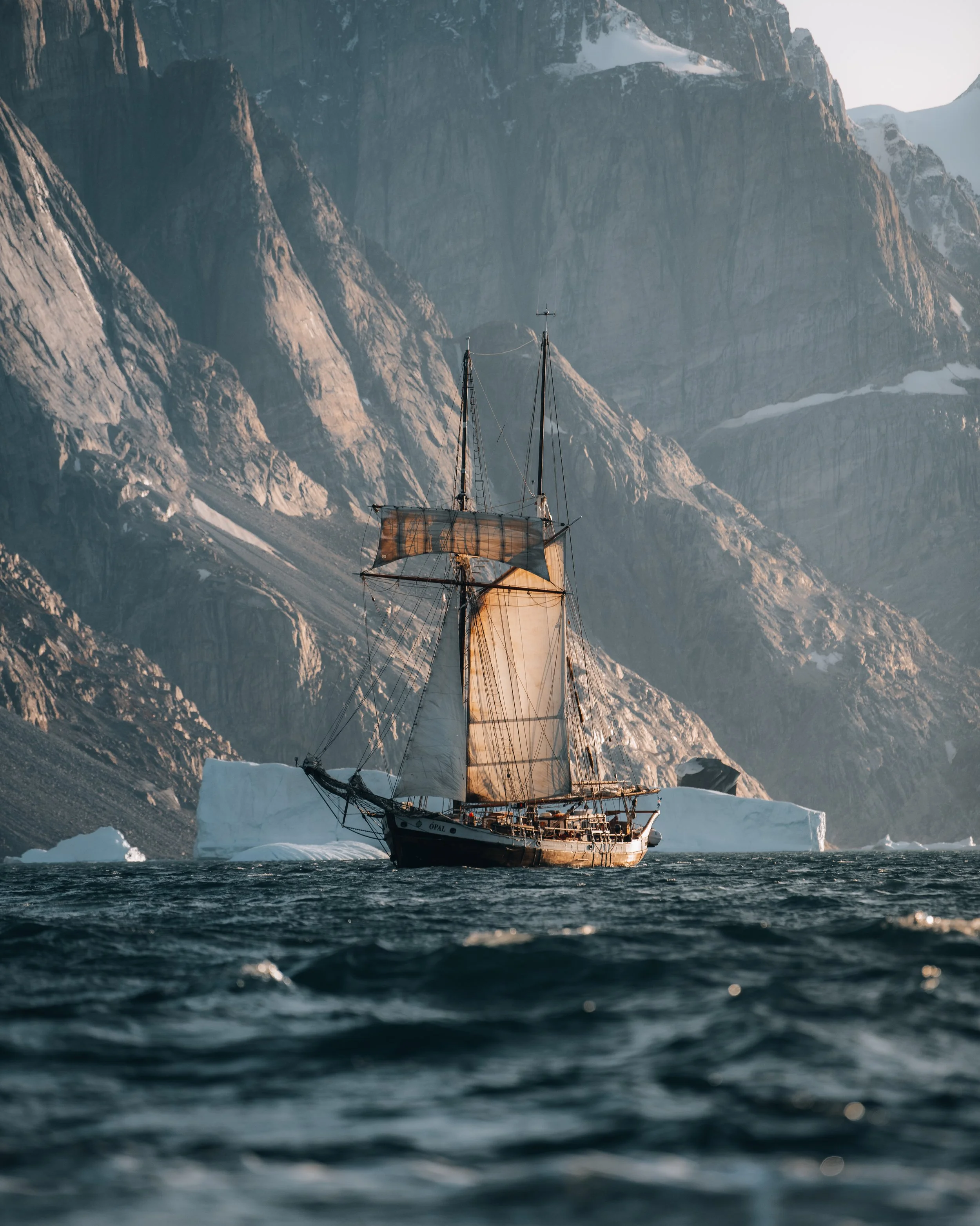 A sailboat navigating icy waters near large icebergs with snow-capped mountains in the background during daylight.