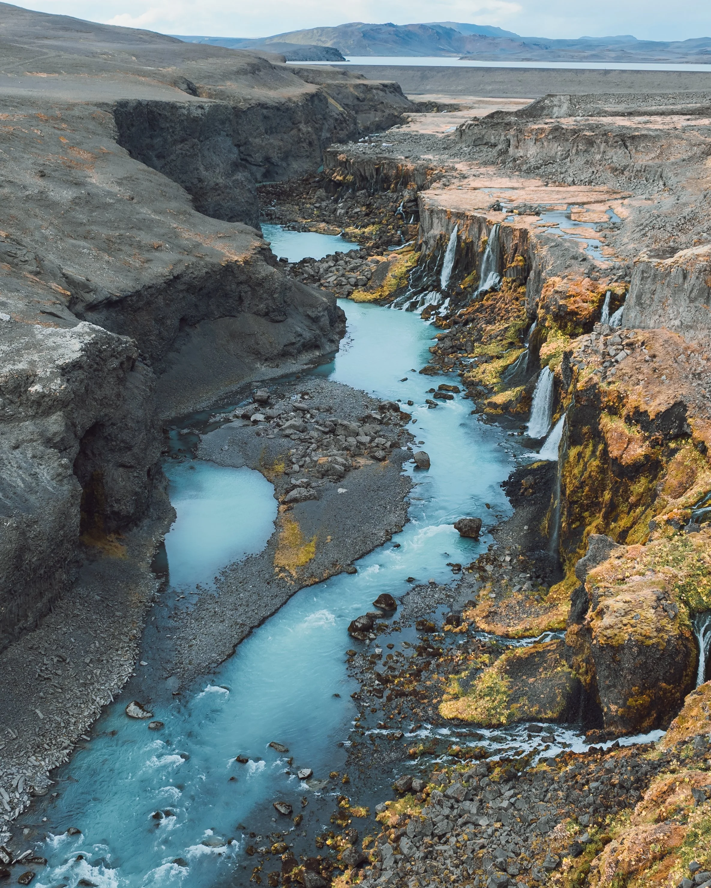 A rugged canyon with a small blue river flowing through it, surrounded by rocky cliffs and volcanic terrain.