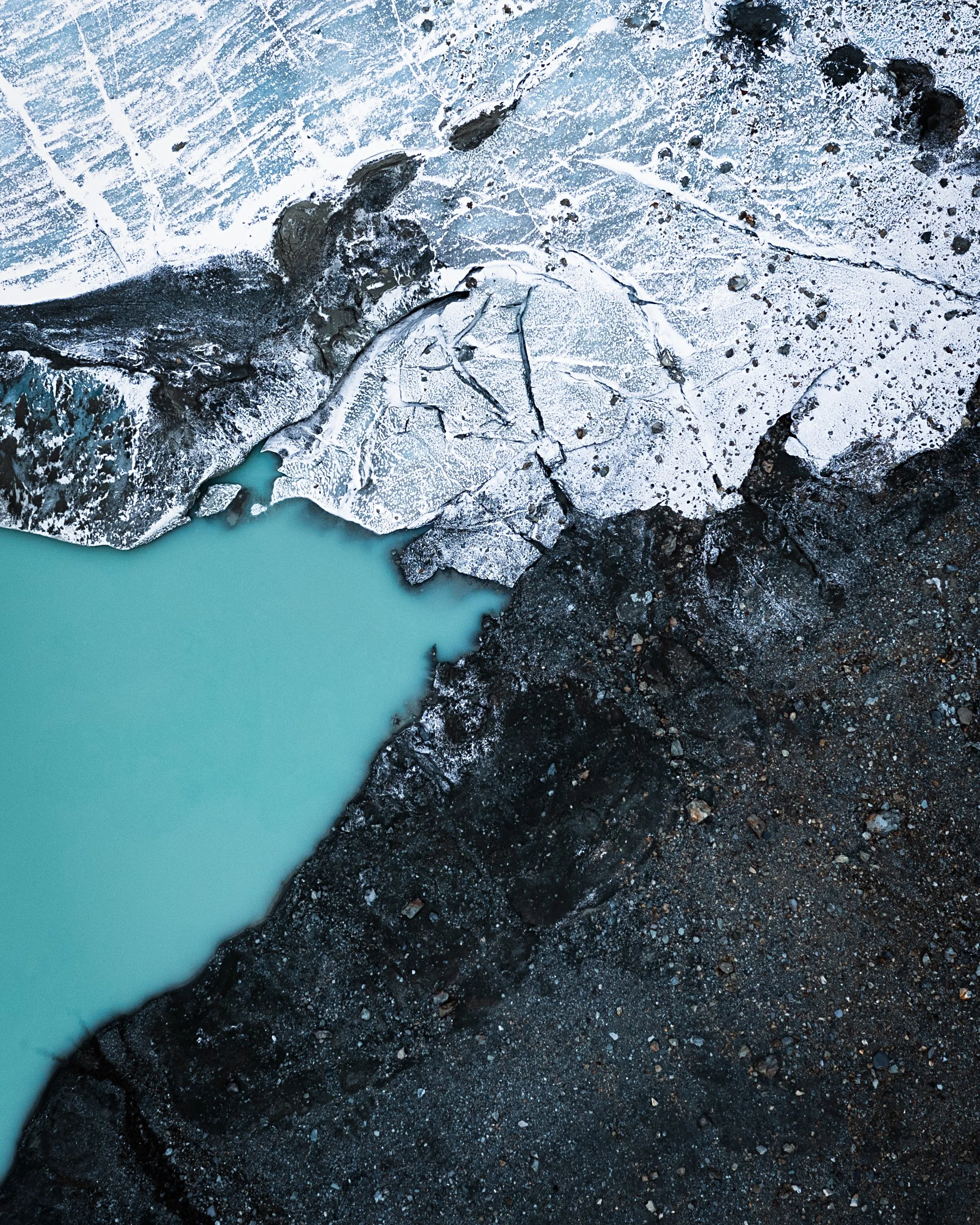 Aerial view of icy glacier meeting a body of milky turquoise water, with dark volcanic rocks surrounding the edges.