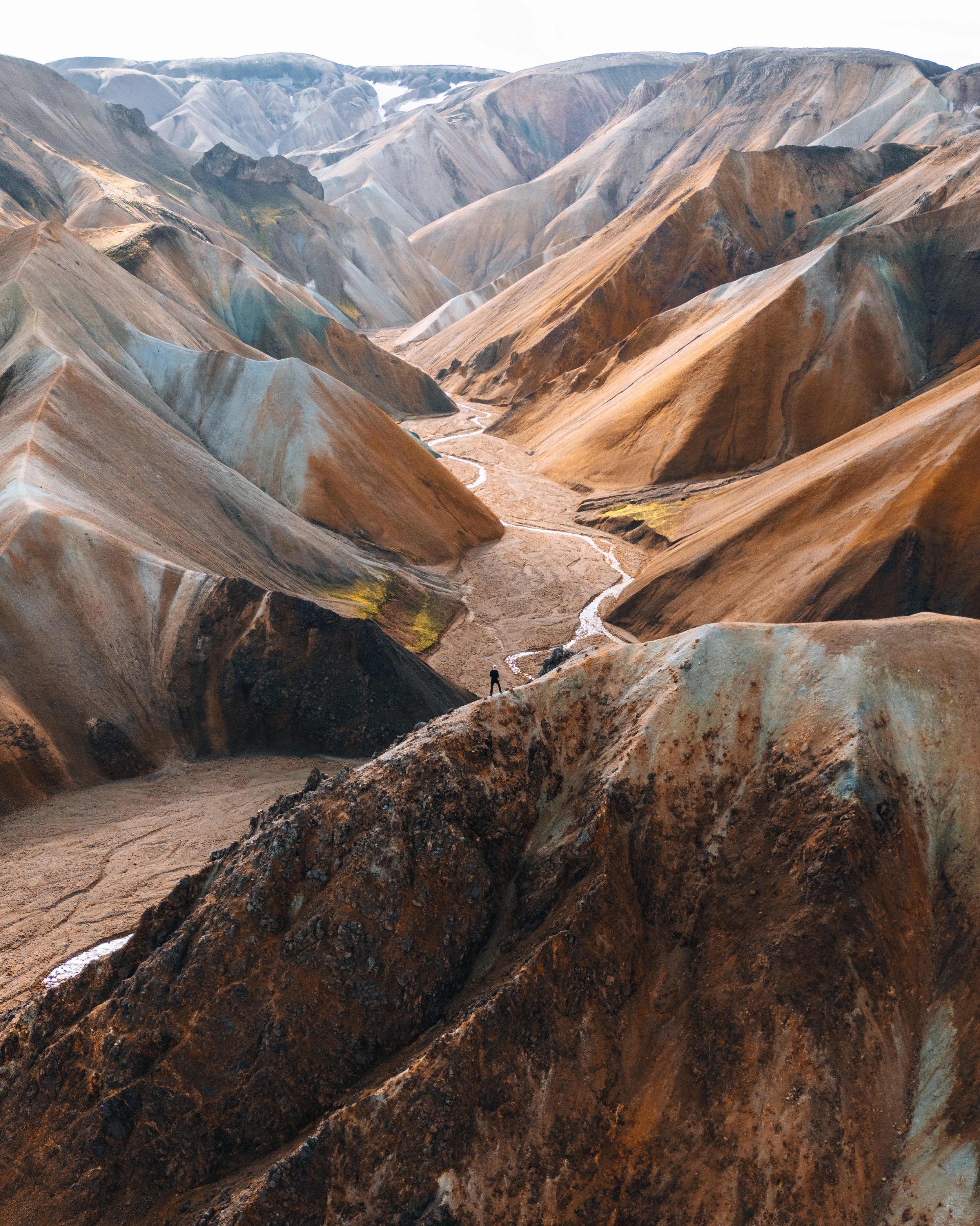 Colorful, layered mountains with a small person standing on the rocky foreground, and a dry riverbed winding through the valley