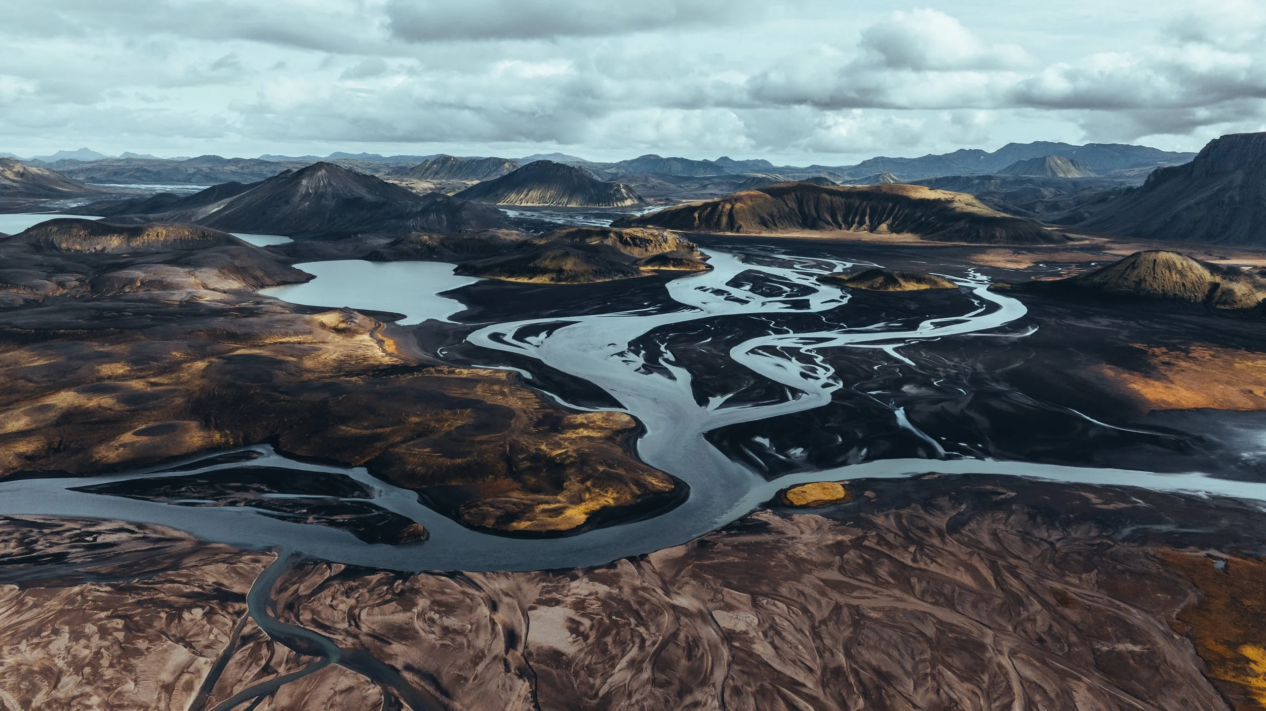 An aerial view of a river winding through a dark, mountainous landscape with hills and mountains in the background under a cloudy sky.
