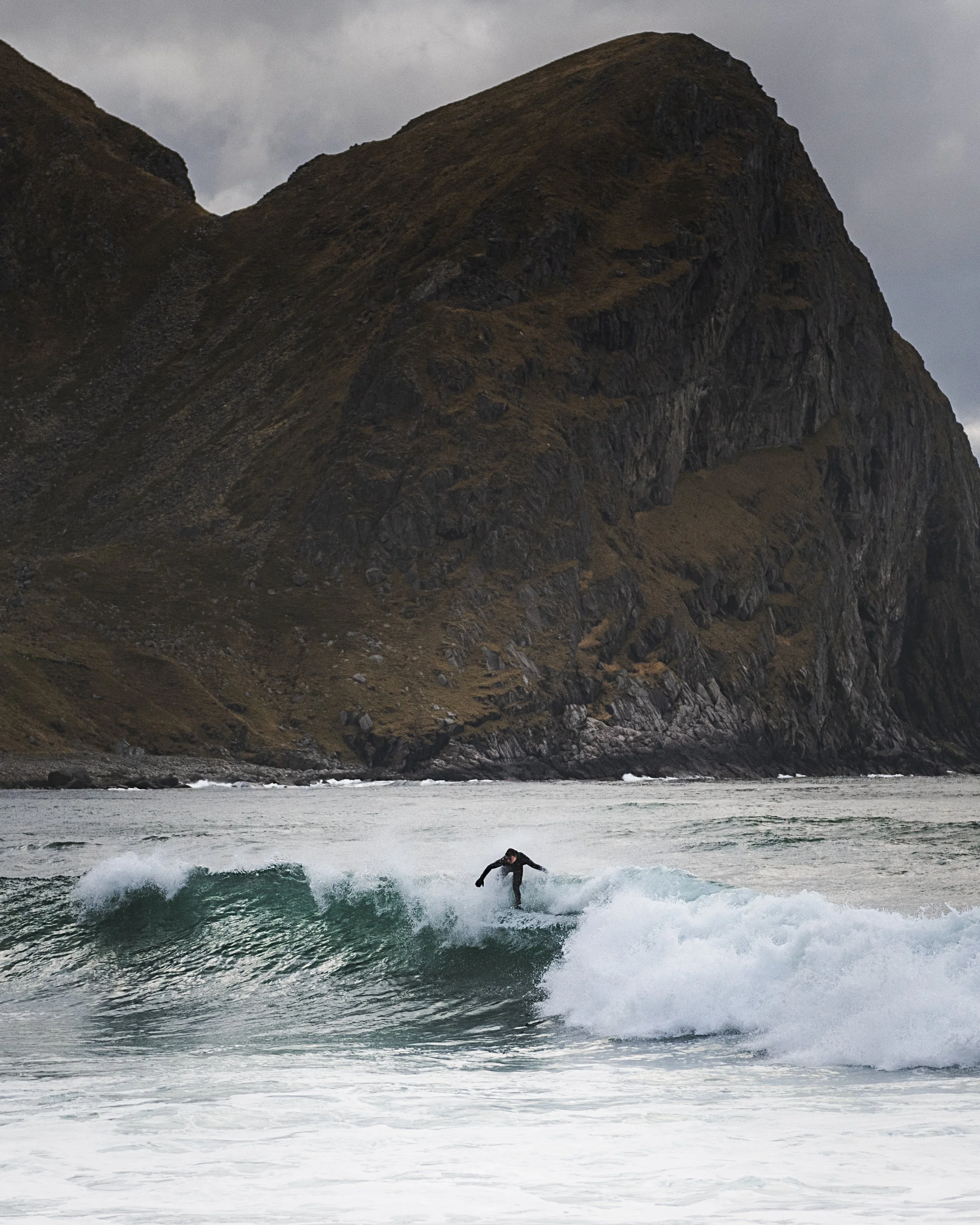 A person surfing on a wave near a rocky coastline with large mountains or cliffs in the background.