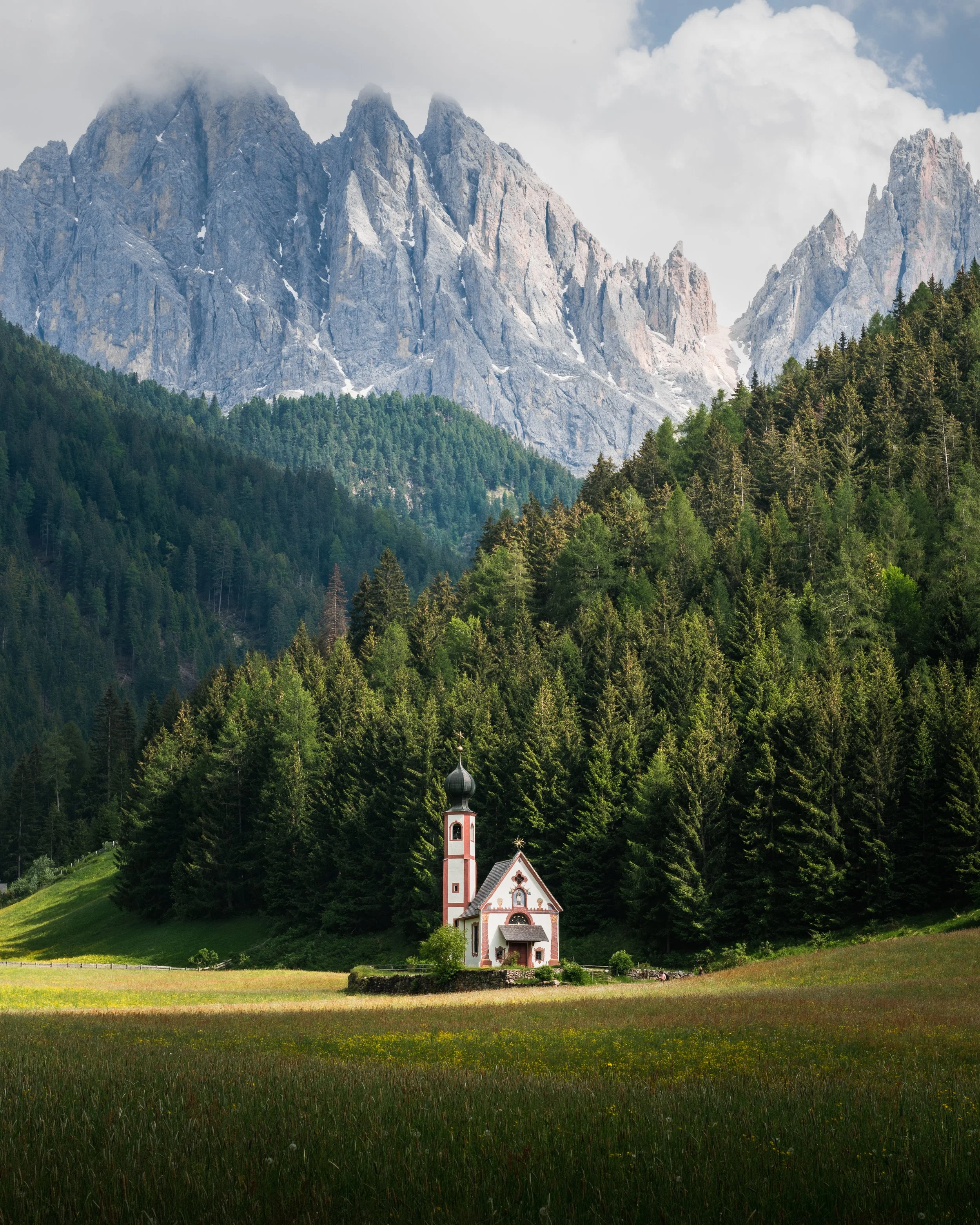 A small white church with a black dome in a meadow surrounded by forest, with mountains in the background.