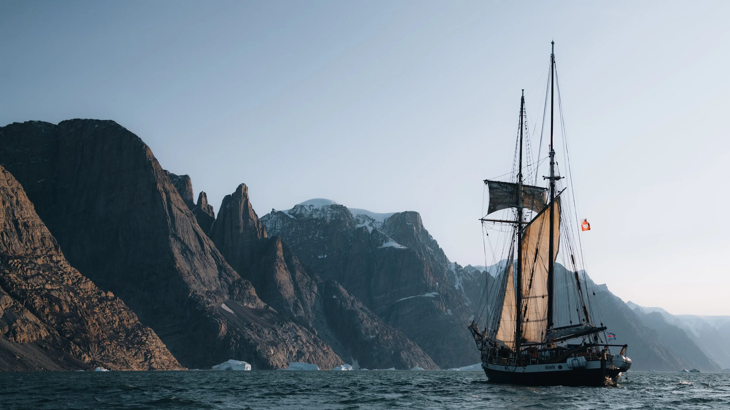 A sailboat sailing near rugged, snow-capped mountains in a body of water during daytime.