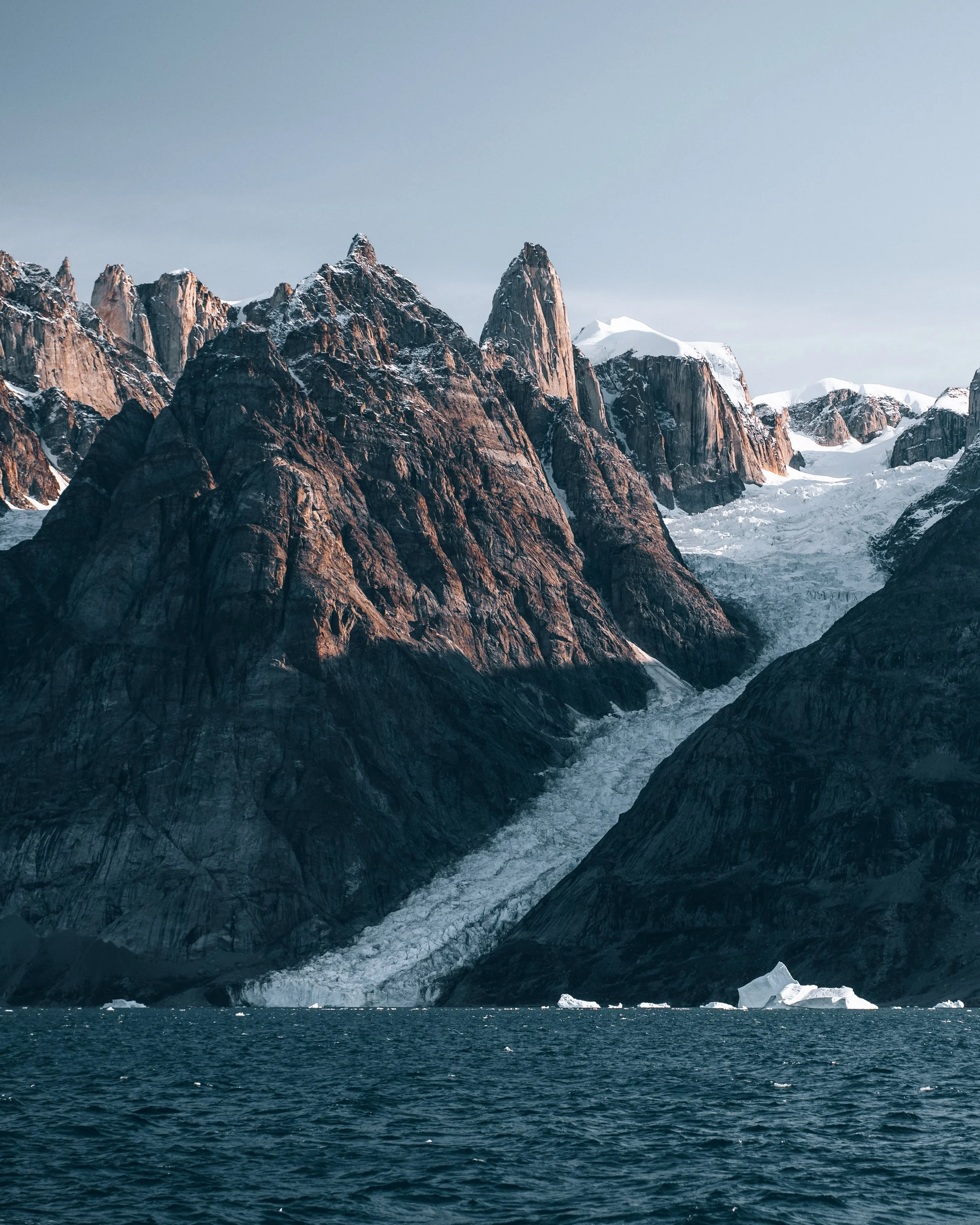 A mountain landscape with tall rocky peaks, snow patches, a glacier, and an ocean with icebergs in the foreground.