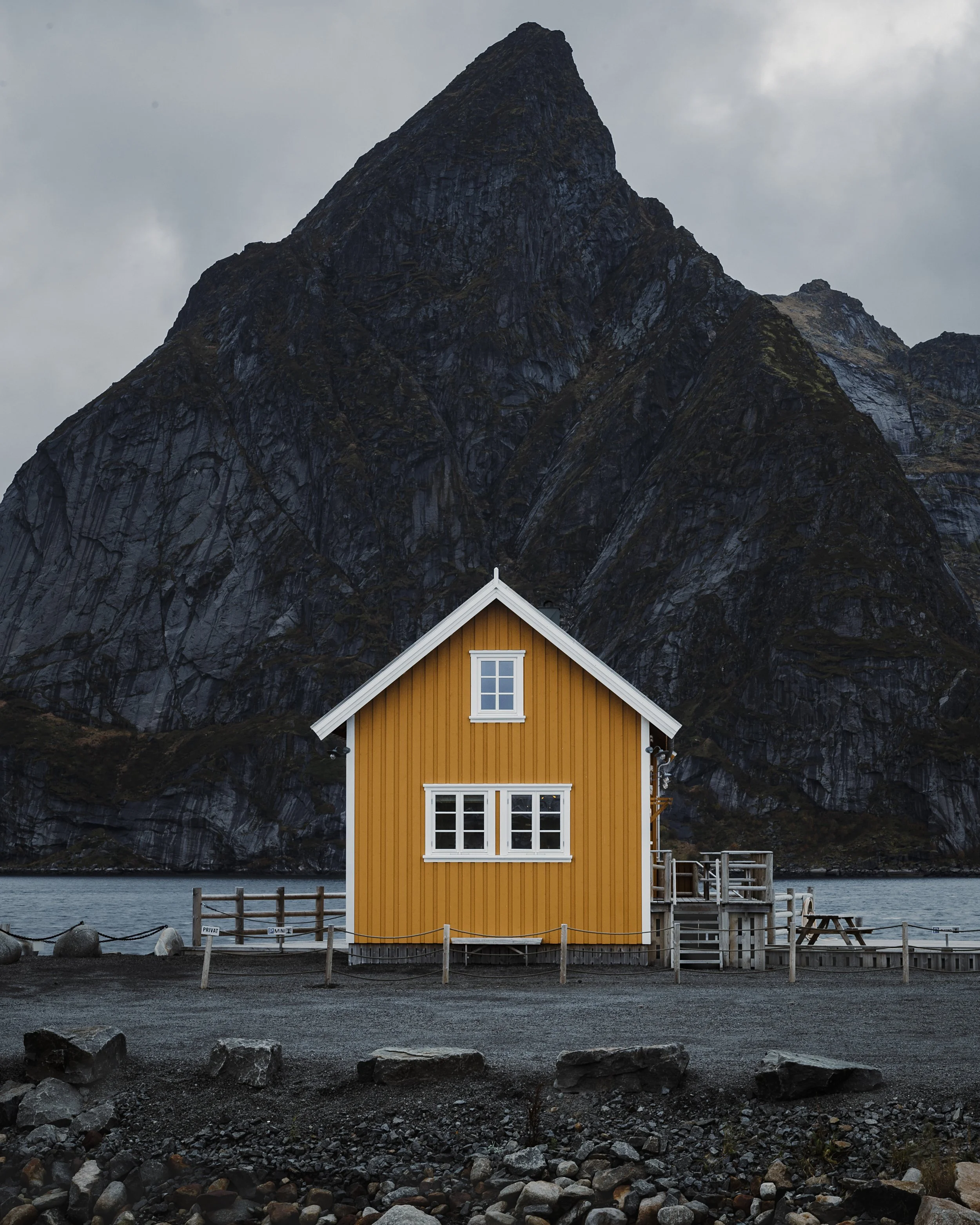 A small yellow house with white windows in front of a large rocky mountain, near a body of water.