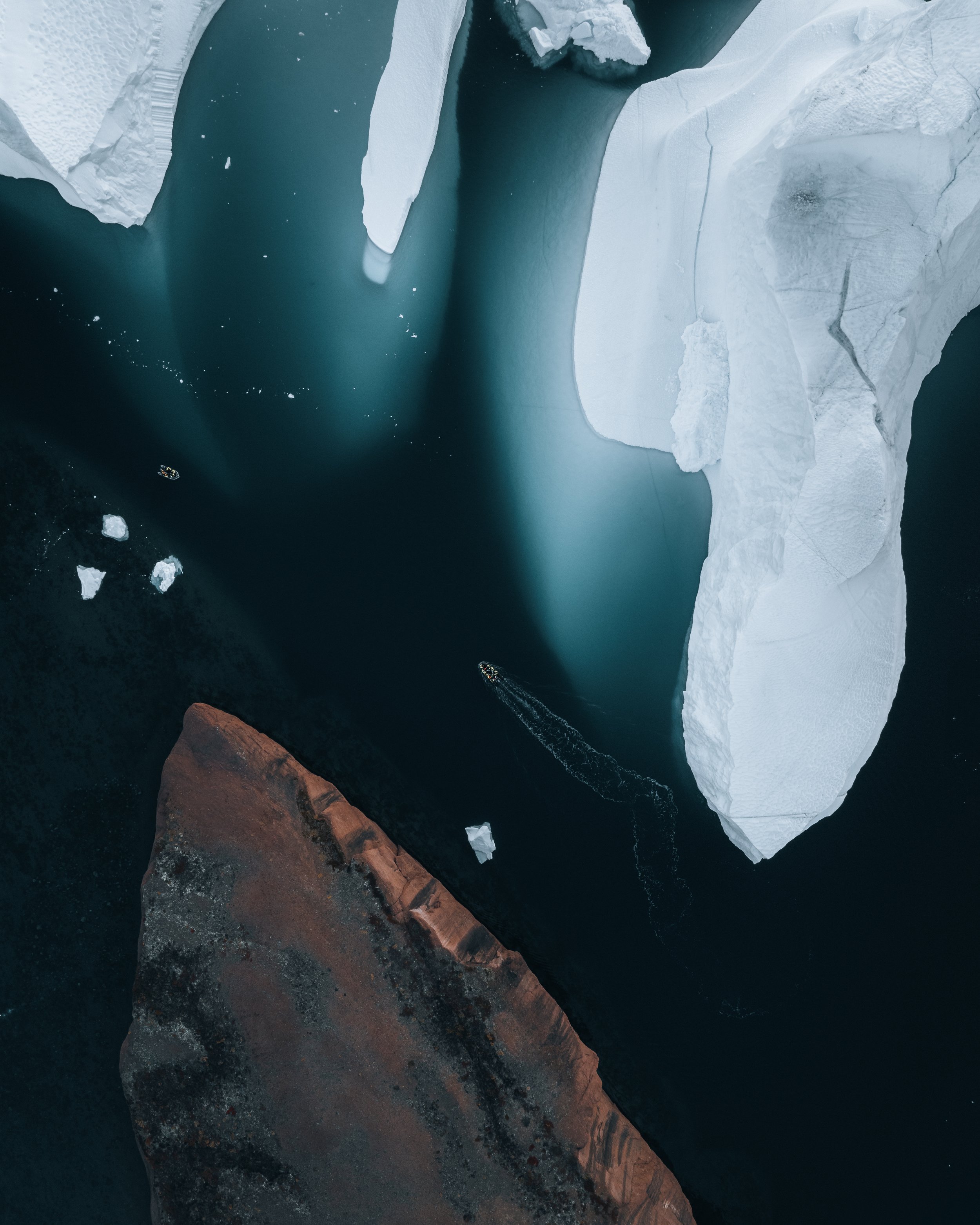 Aerial view of icy landscape with large white ice formations, a dark blue water body, and a small boat leaving a trail in the water.