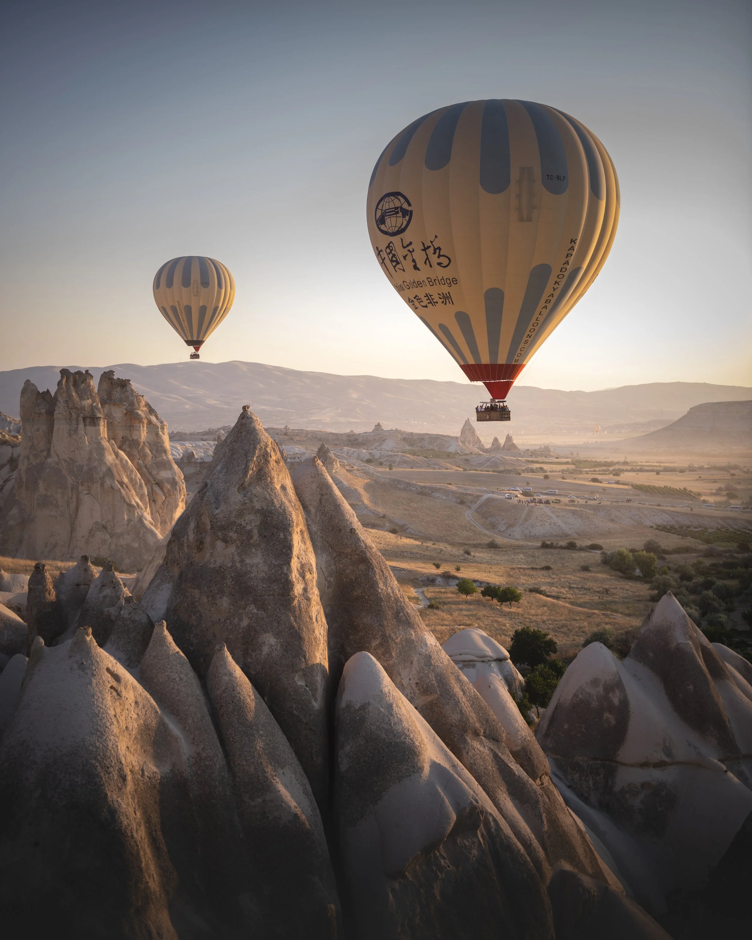 Two hot air balloons floating over rocky landscape during sunset, with valleys and distant mountains in the background.