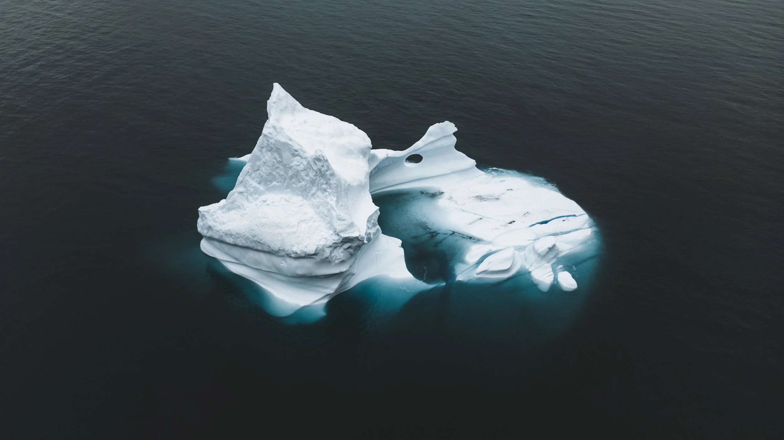 An iceberg floating in dark ocean water, with a sharp peak and a hole in one of the ice formations.