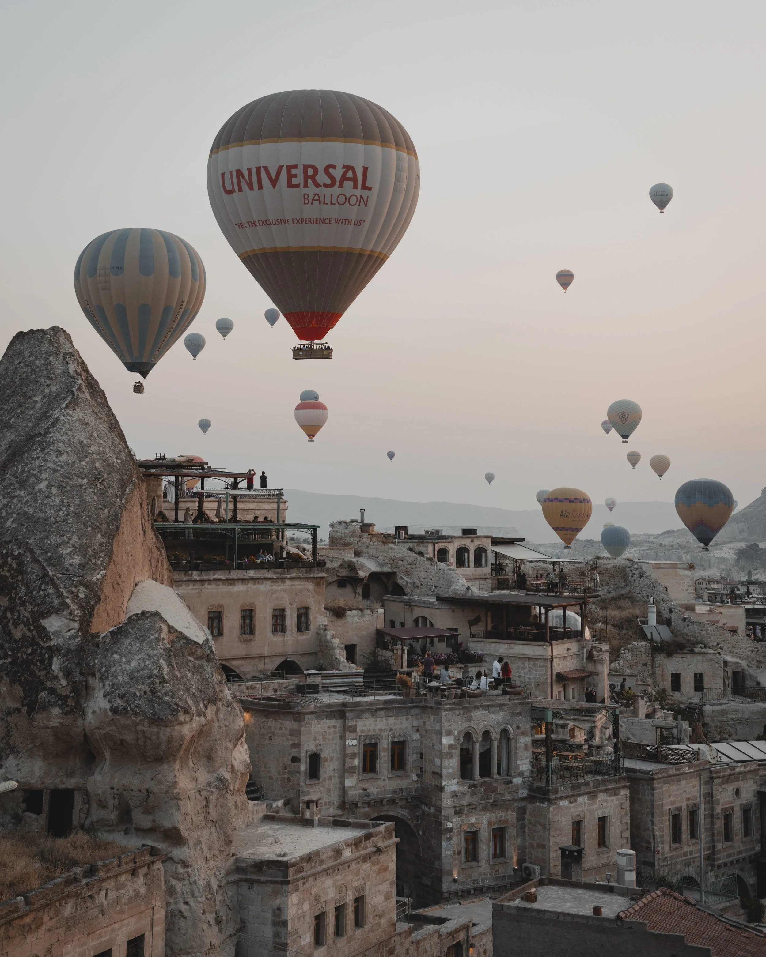 Multiple hot air balloons floating over an ancient city with stone buildings and rock formations during sunrise or sunset.