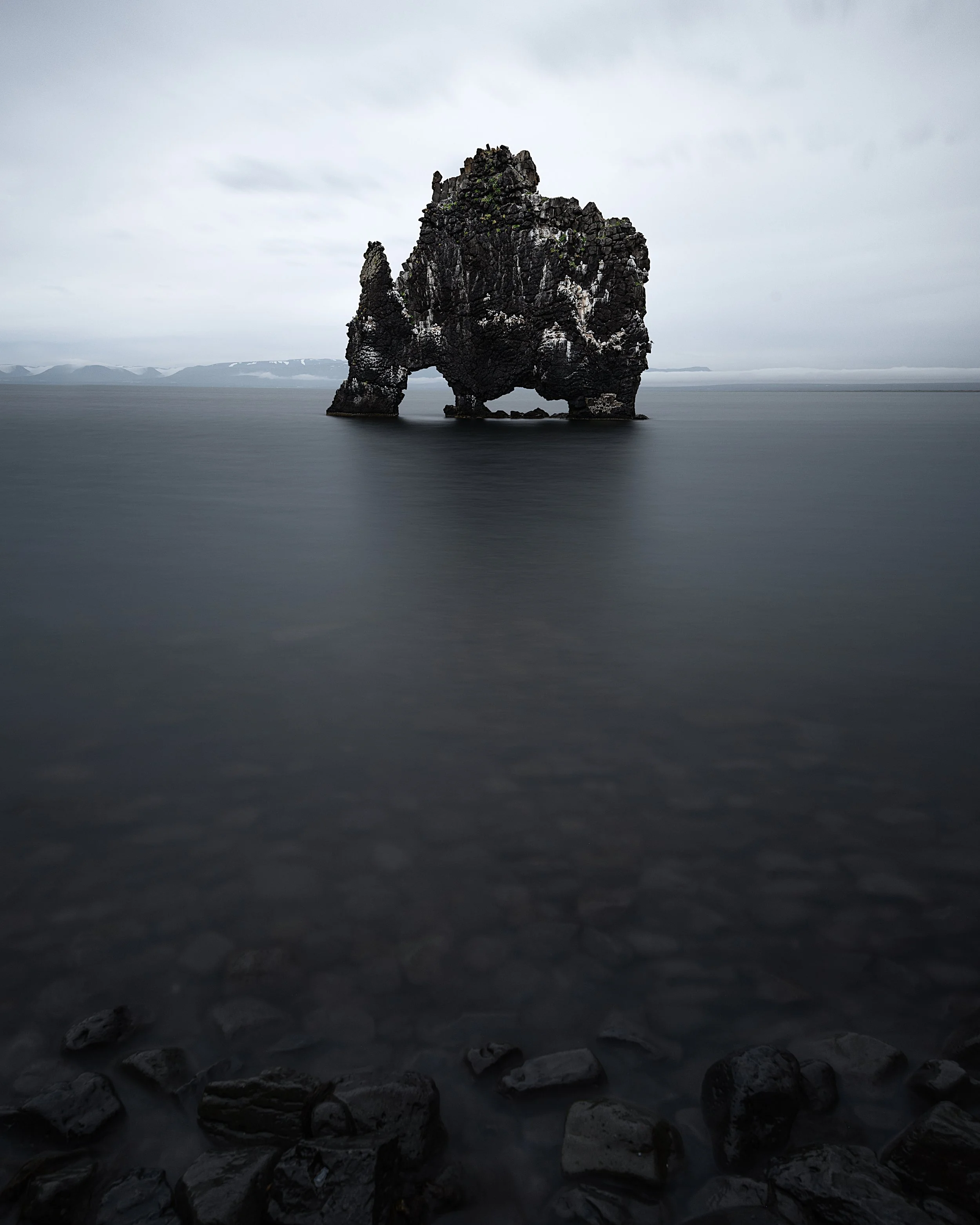 A large rock formation with a natural arch near the water's surface in a gray, overcast landscape