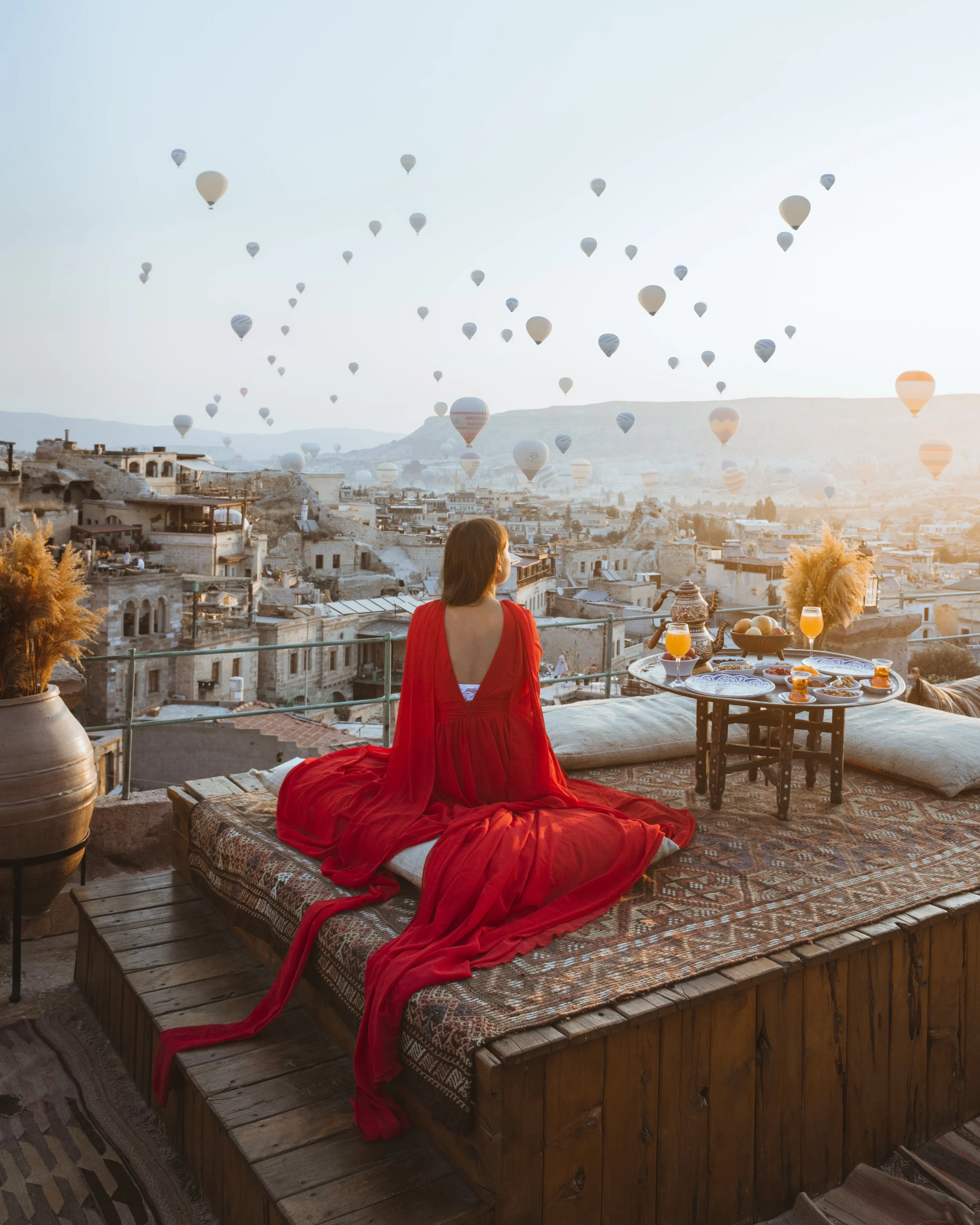 A woman in a red dress sitting on a rooftop lounge, overlooking a cityscape with numerous hot air balloons in the sky at sunrise or sunset.
