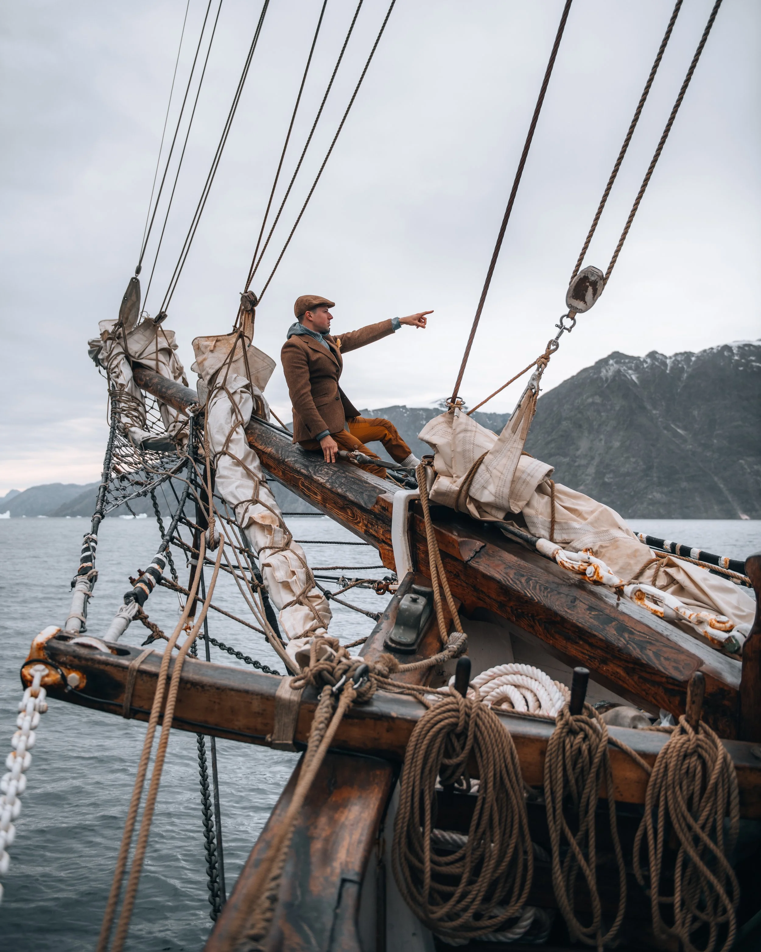 A man sitting on the wooden deck of a sailing ship, pointing forward, with mountains in the background and the sea below.