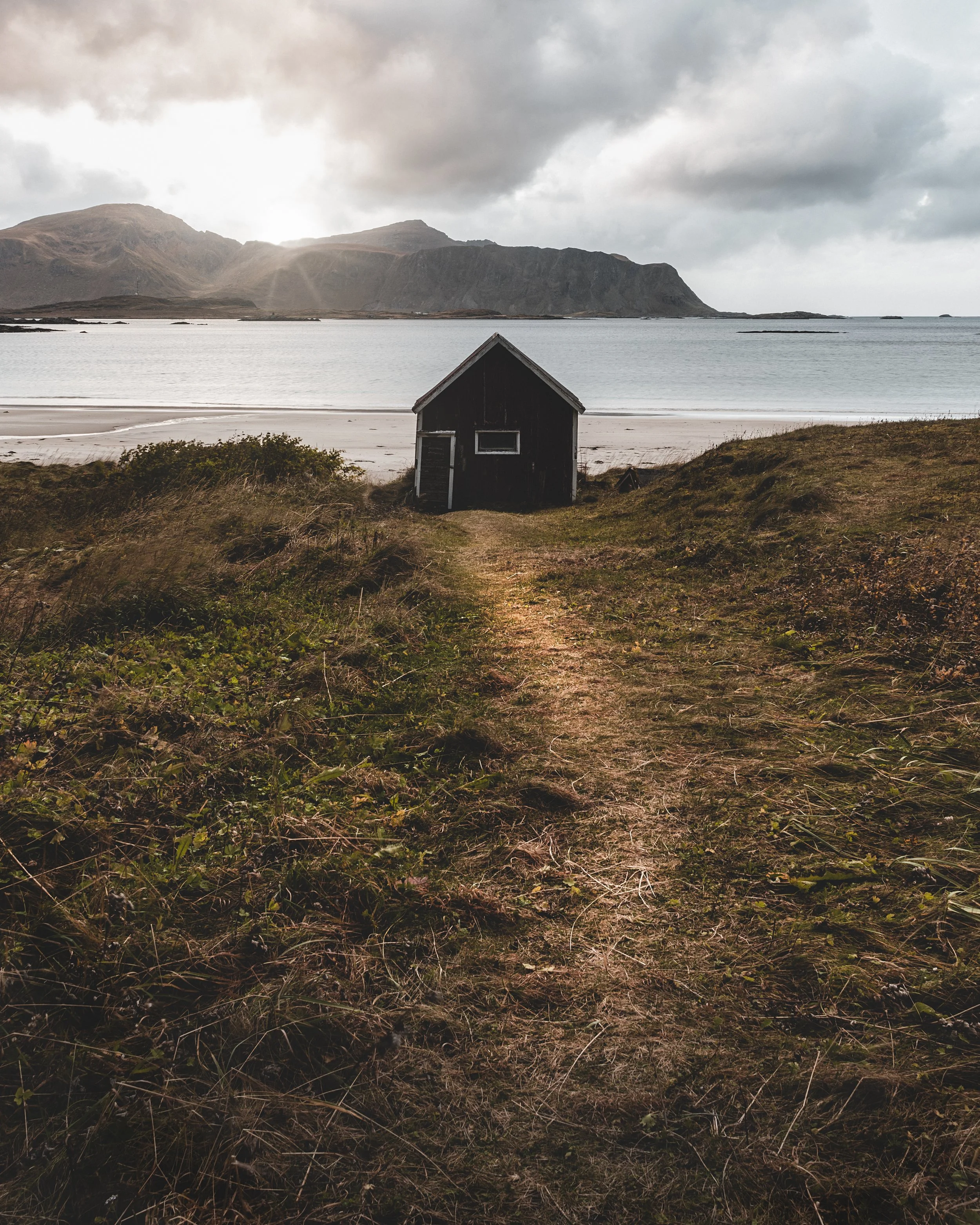 A narrow dirt path leading to a small black wooden house on a beach with mountains in the background and a cloudy sky.