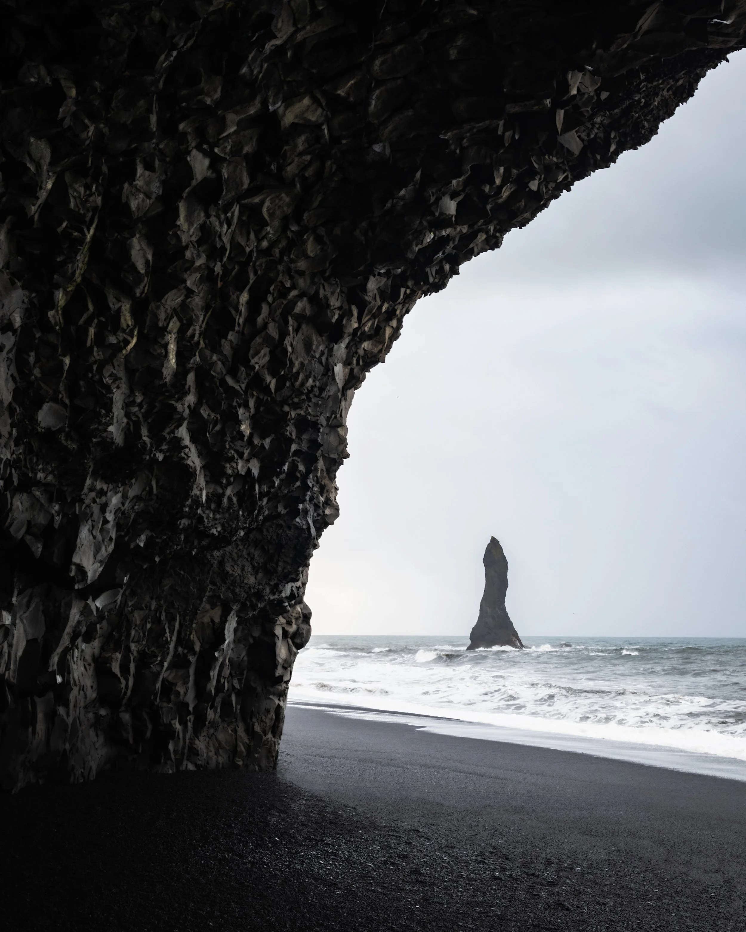 A dark cave opening on a black sand beach with a large sea stack rock formation in the ocean in the background under an overcast sky.