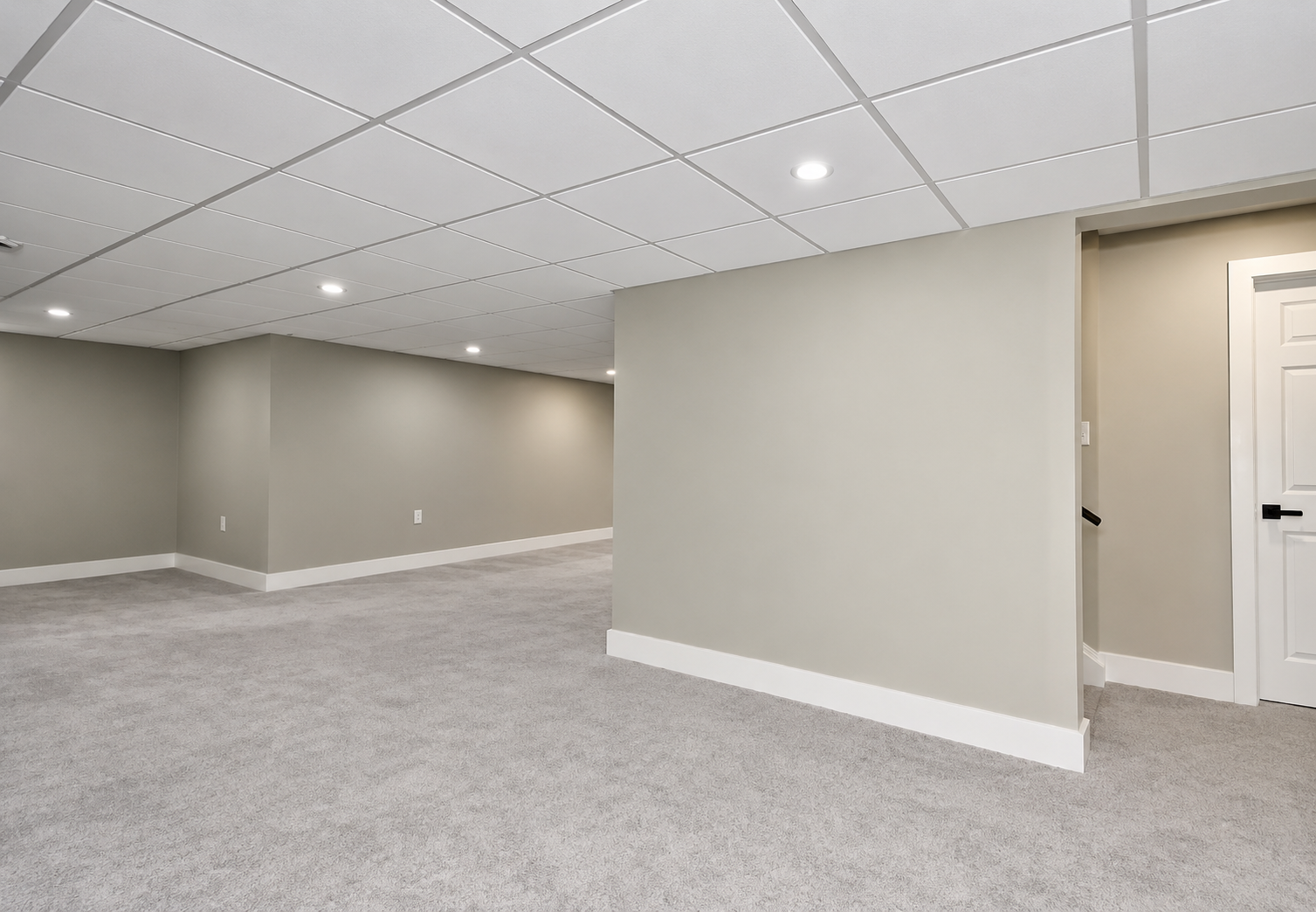 Empty basement room with light gray walls, white baseboards, gray carpeting, white ceiling tiles, and recessed lighting, with a doorway on the right side.