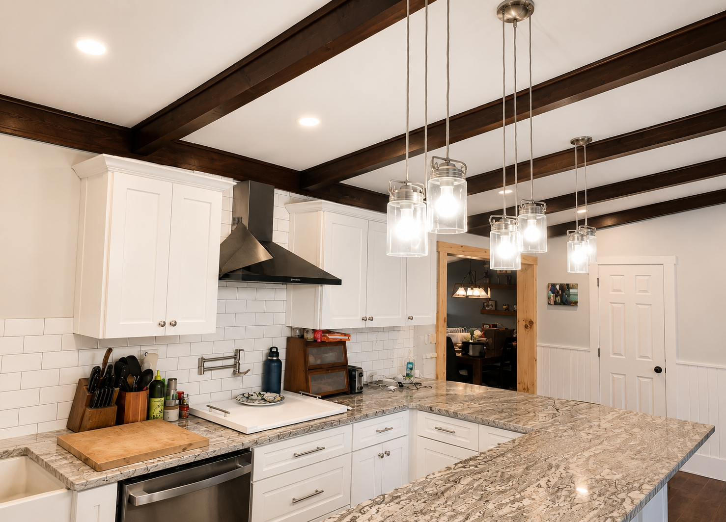 Modern kitchen with white cabinets, granite countertops, and dark wooden ceiling beams. A black range hood is above the stove, and a hanging light fixture with multiple bulbs is centered over the kitchen island.