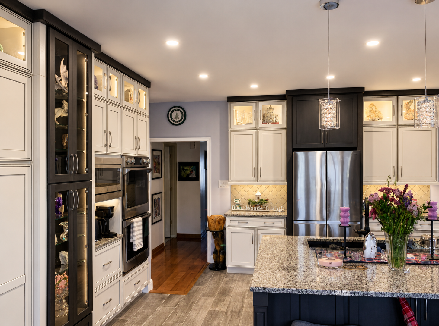 Modern kitchen with white and black cabinets, stainless steel refrigerator, granite countertops, and decorative glass display cabinets. There are pink candles, a bouquet of purple flowers, and pendant lights overhead.