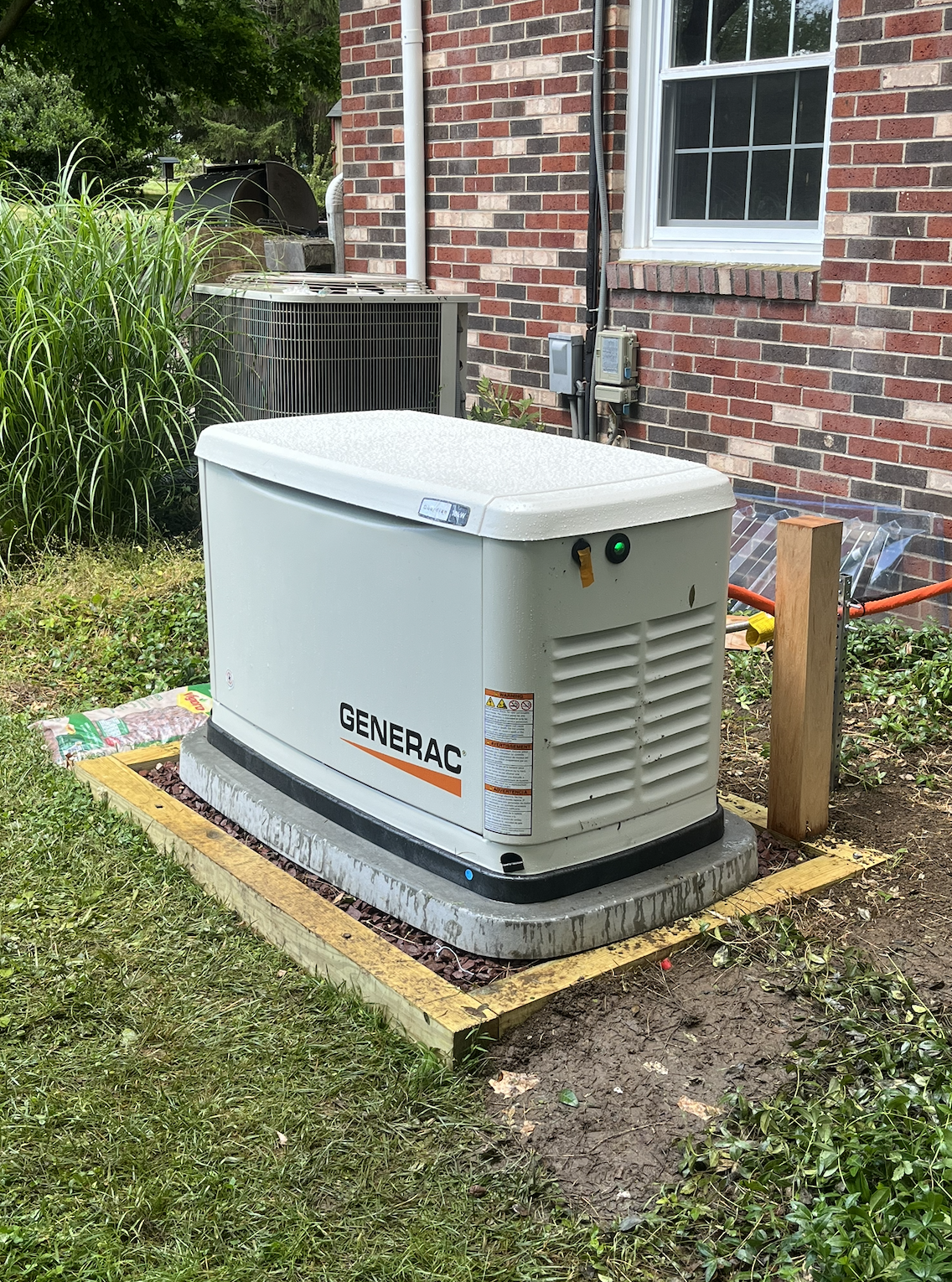 A Generac standby generator installed on a concrete slab outside a house, surrounded by grass and nearby plants, with a brick house wall, a window, and HVAC unit in the background.
