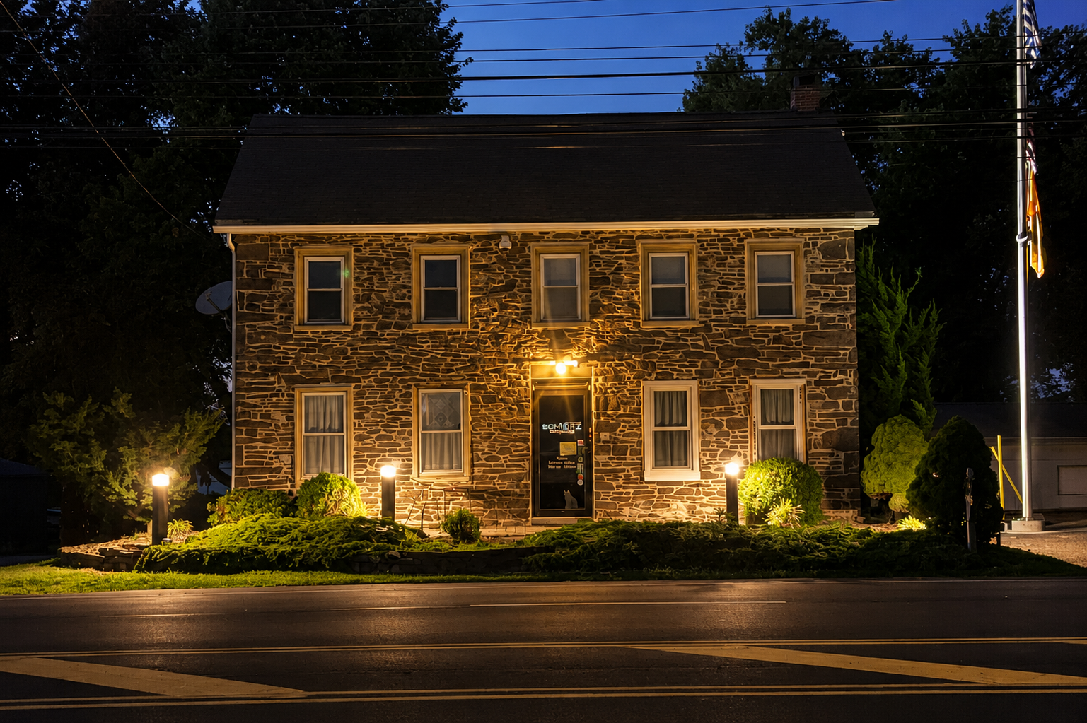 A two-story brick building with six windows and a door at night, illuminated by exterior lights, with a lawn and shrubbery in front and trees in the background.