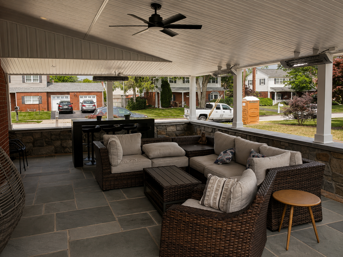 Covered porch with wicker furniture, cushioned sofas, and pillows overlooking a suburban neighborhood street with cars, houses, trees, and a portable toilet.
