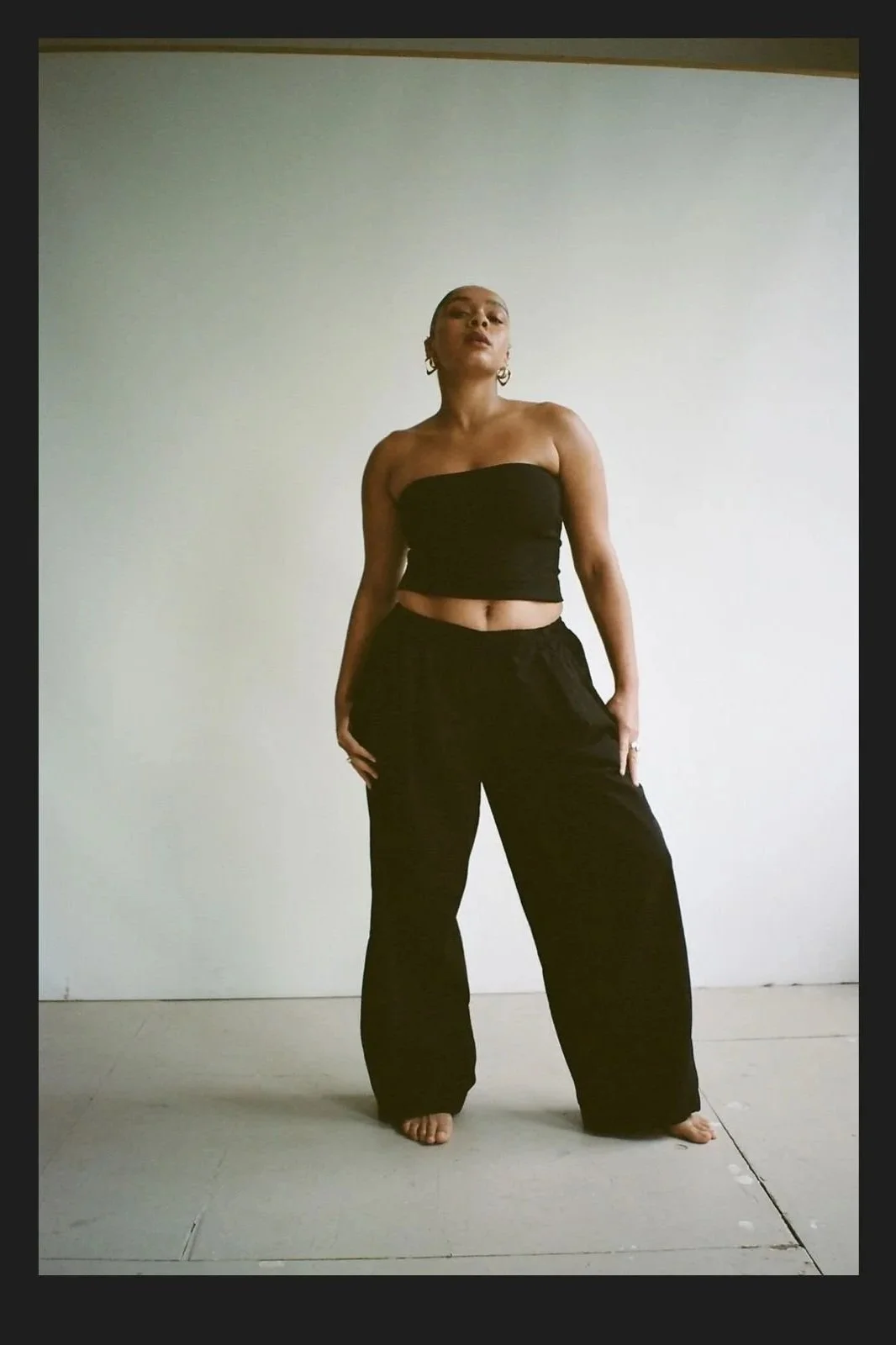 A woman stands barefoot against a plain white wall, wearing a black tube top and loose black pants, with her hands resting on her hips.