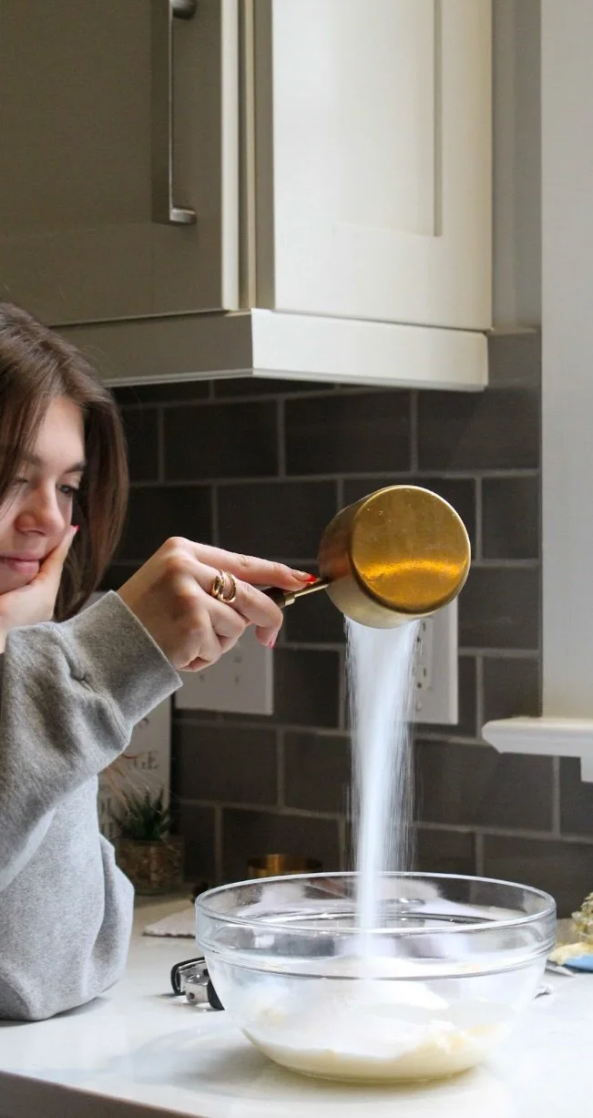 A person pouring sugar from a copper measuring cup into a glass bowl on a kitchen counter.