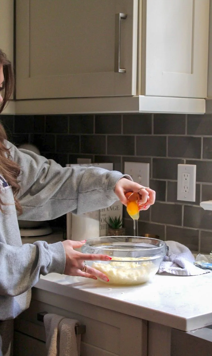 Person cracking an egg into a glass bowl on a kitchen counter with gray tile backsplash.
