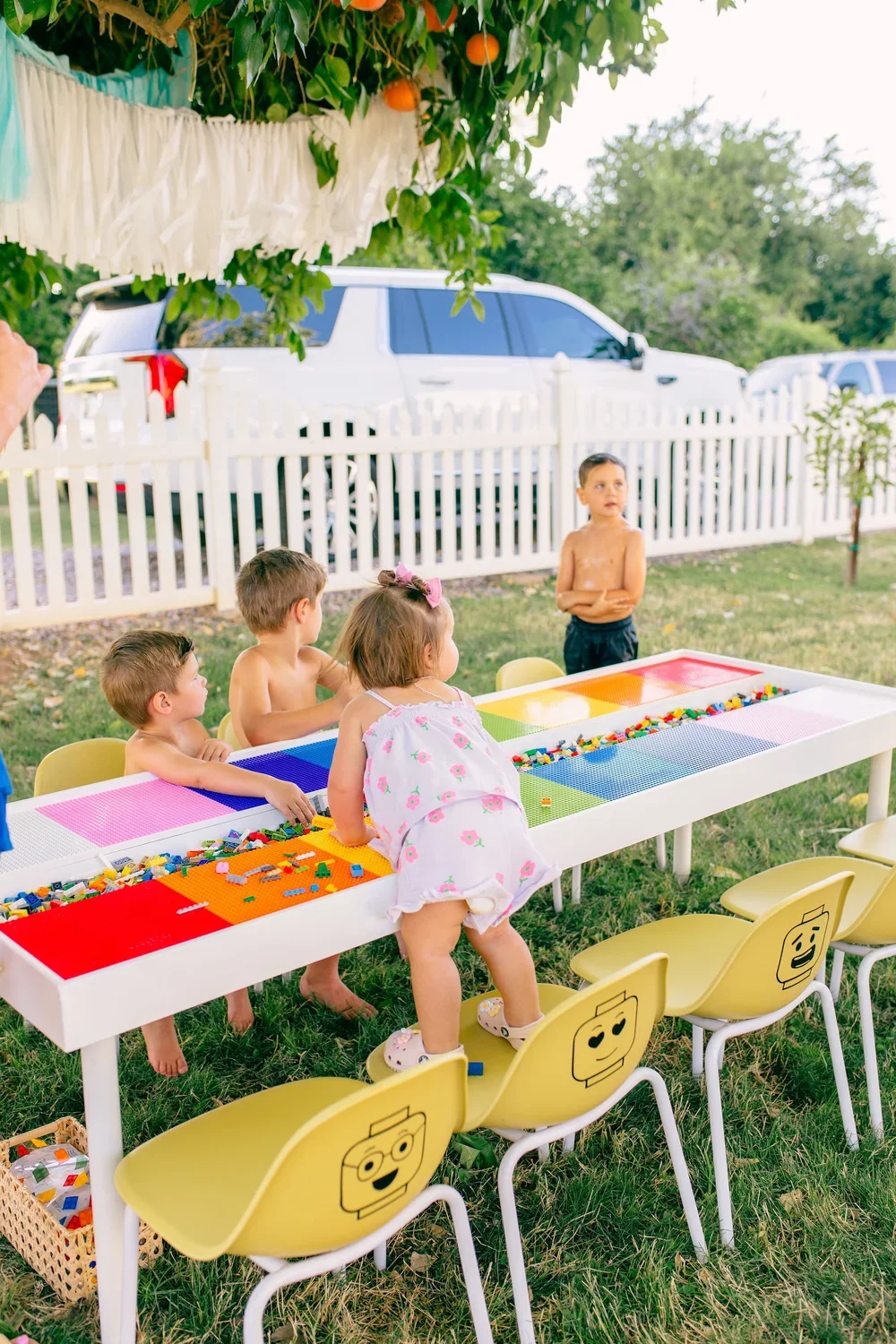 Children playing with LEGO bricks at a colorful outdoor table during a backyard gathering, with four children standing and one child sitting, beside a white picket fence and parked cars in the background.