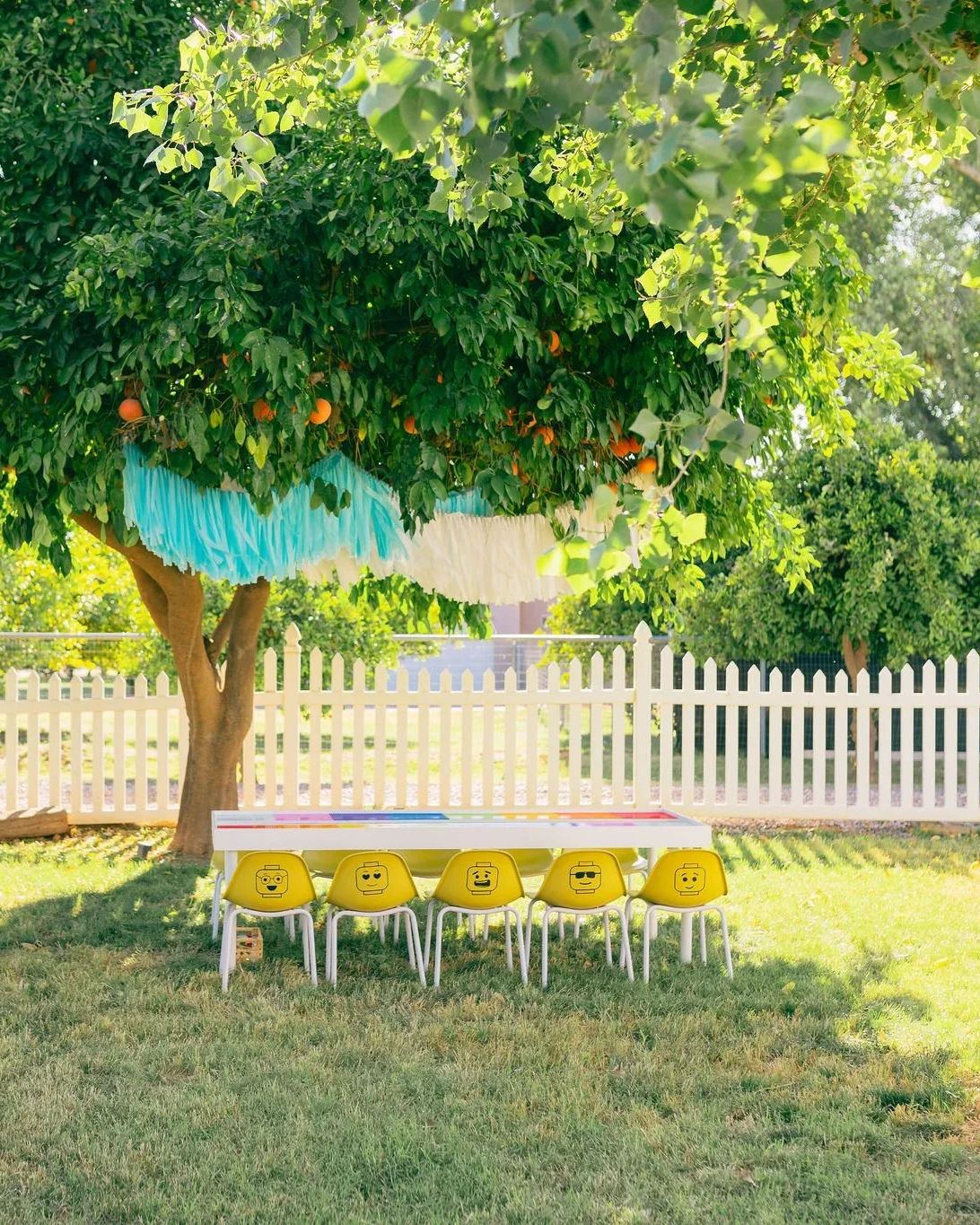 Outdoor scene with a large green tree decorated with colorful hanging paper garland and orange fruit. Under the tree, there is a white table with colorful markings and five yellow chairs, each featuring a different emoji face. A white picket fence surrounds the area on a sunny day.
