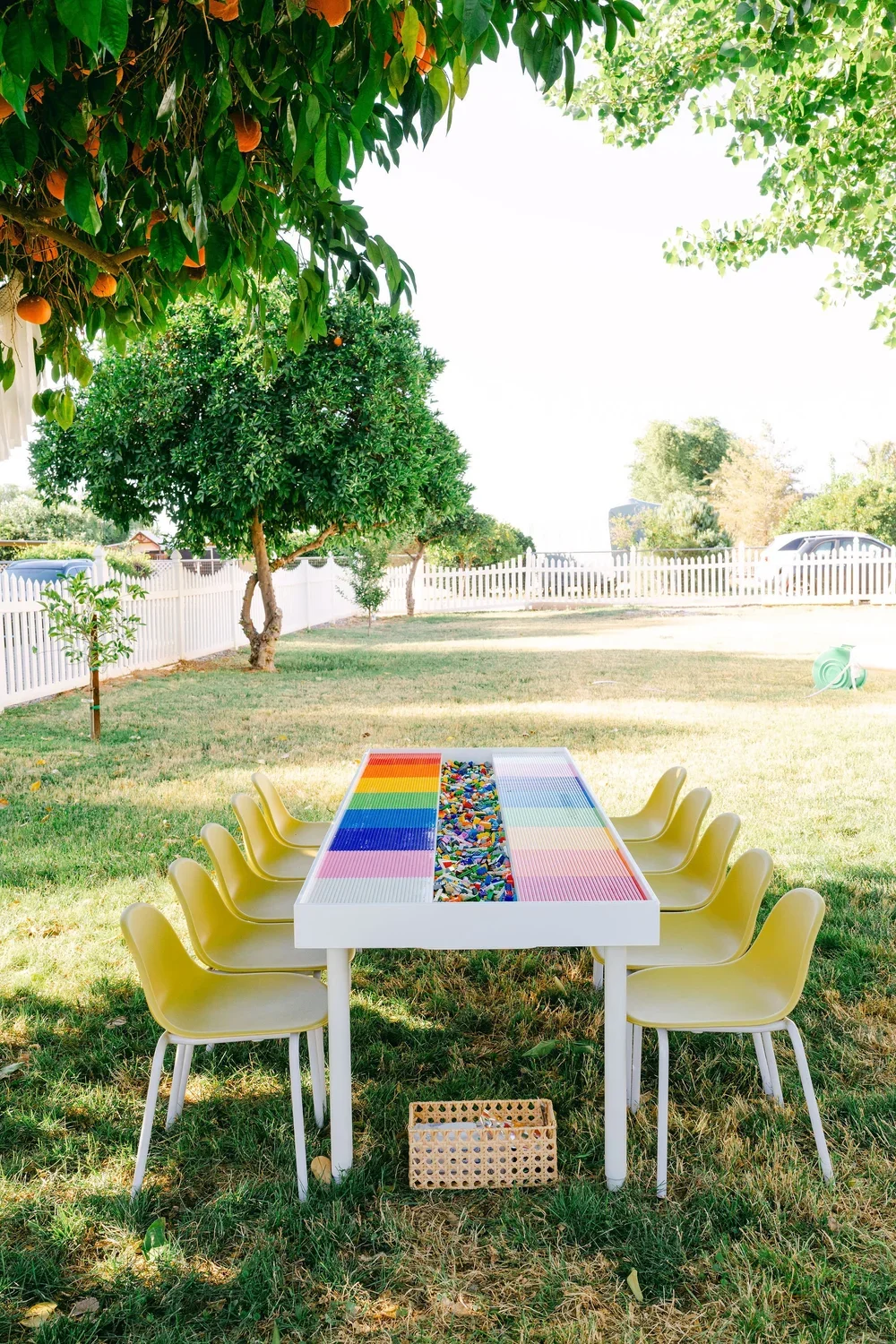 Outdoor scene with a white Lego table surrounded by eight yellow chairs, a small basket underneath, and trees with orange fruits and a white picket fence in the background.