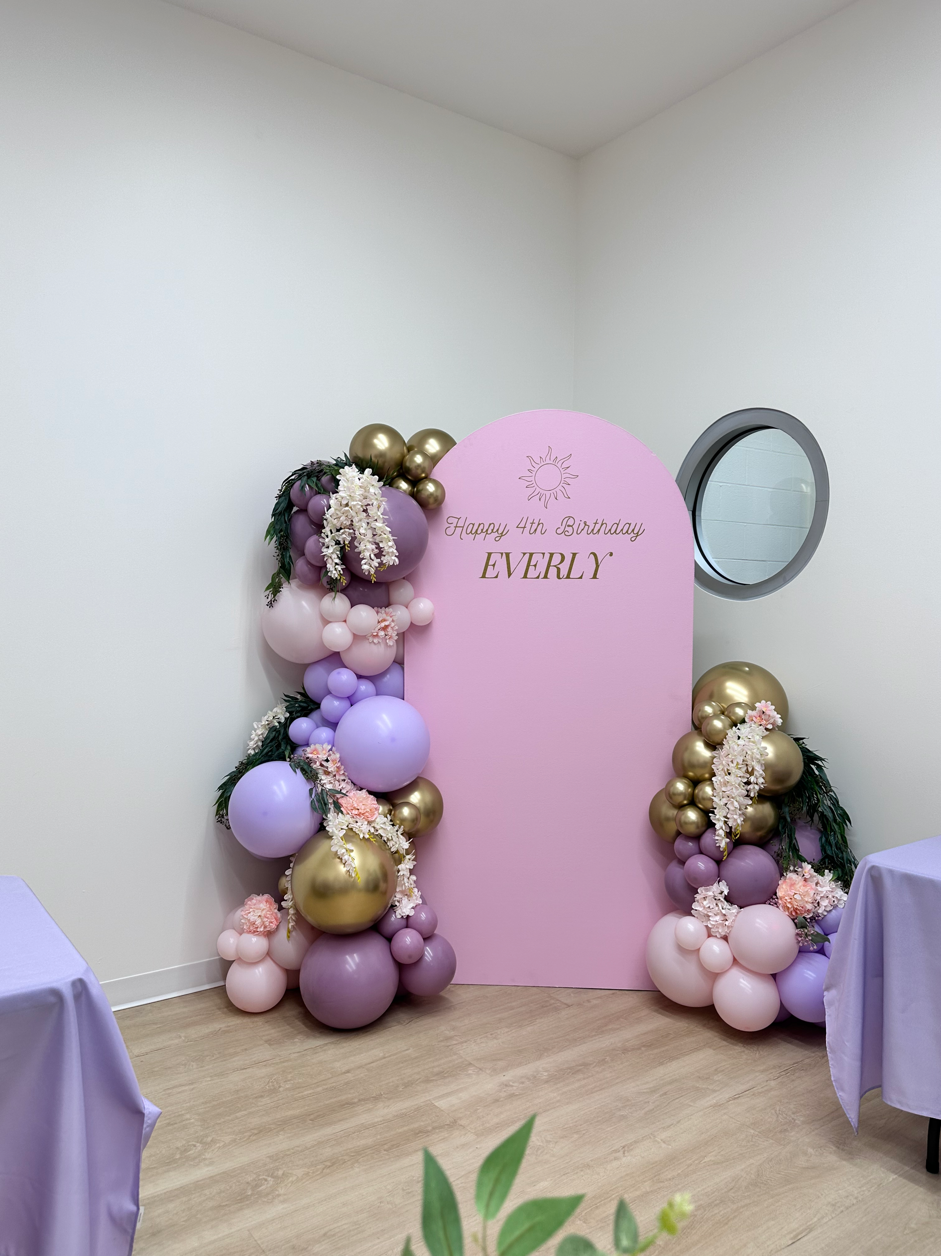 Pink backdrop with birthday message and balloon decorations for a 4th birthday celebration, featuring purple, gold, and pink balloons with flowers.