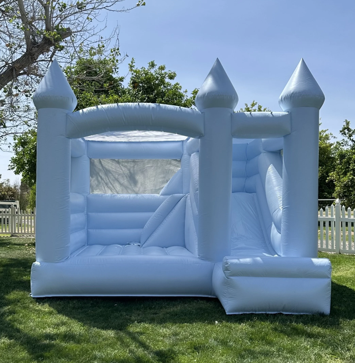 White inflatable bounce house resembling a castle with four turrets, set up on a grassy area outdoors near a white picket fence and trees.