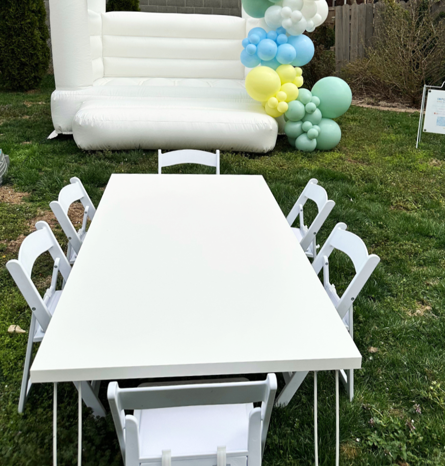 An outdoor setup with a white table and chairs on green grass, decorated with pastel-colored balloons, and a white inflatable structure in the background.