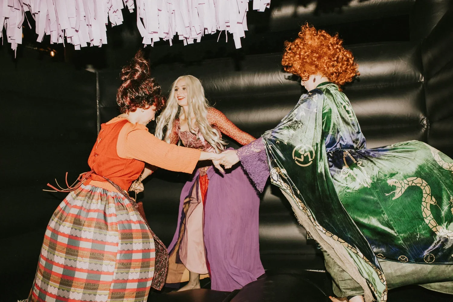 Three women dressed in colorful, elaborate costumes holding hands and smiling on a dark stage decorated with pink paper strips hanging from the ceiling.