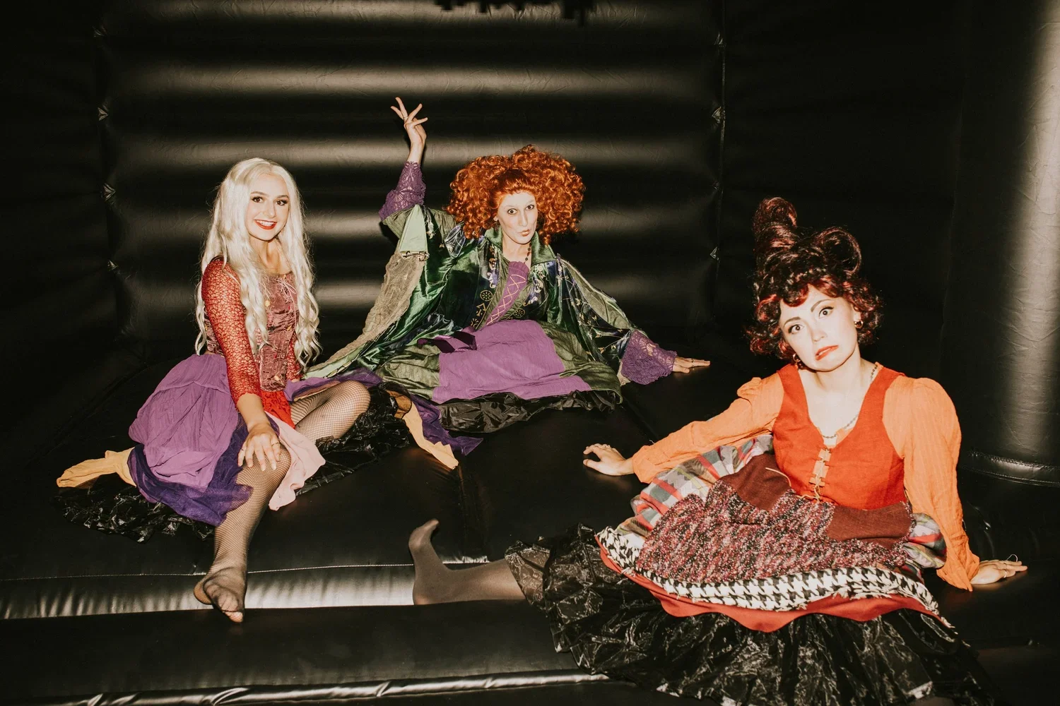 Three women dressed in colorful, theatrical costumes sitting on a black inflatable surface inside a black inflatable enclosure.
