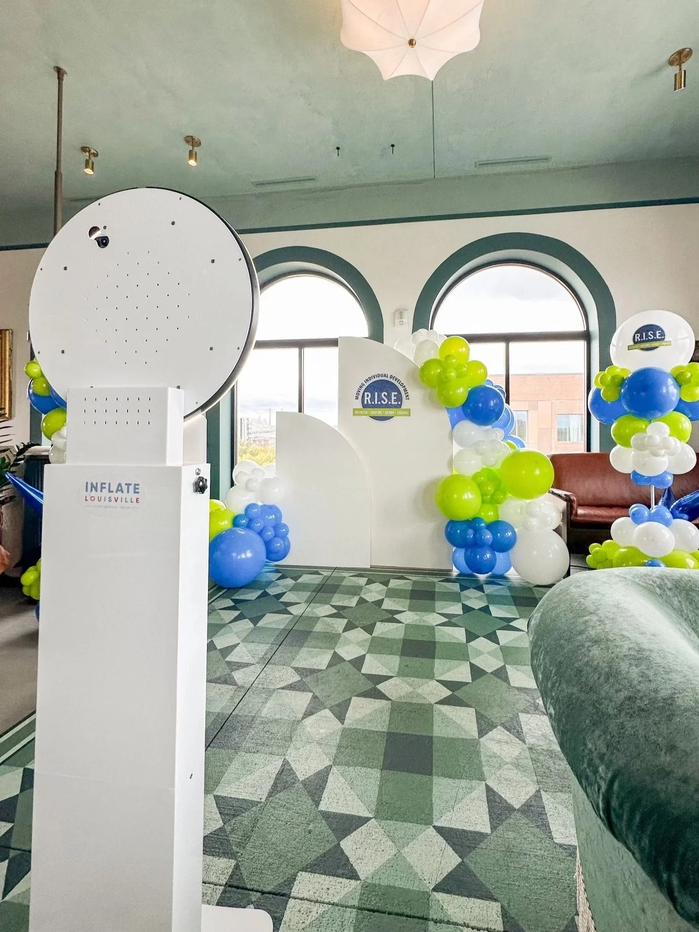Decorative area with balloon arrangements in white, blue, and green around a white backdrop with seating, near arched windows, with a patterned green and gray floor.