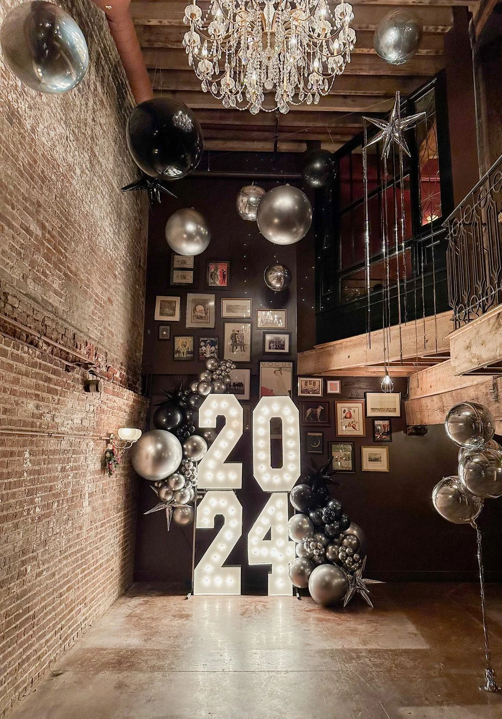 Decorations for a 2024 celebration featuring large illuminated numbers '20' and '24' with balloons and framed pictures on the wall in a dimly lit room with exposed brick wall, chandelier, and string lights.