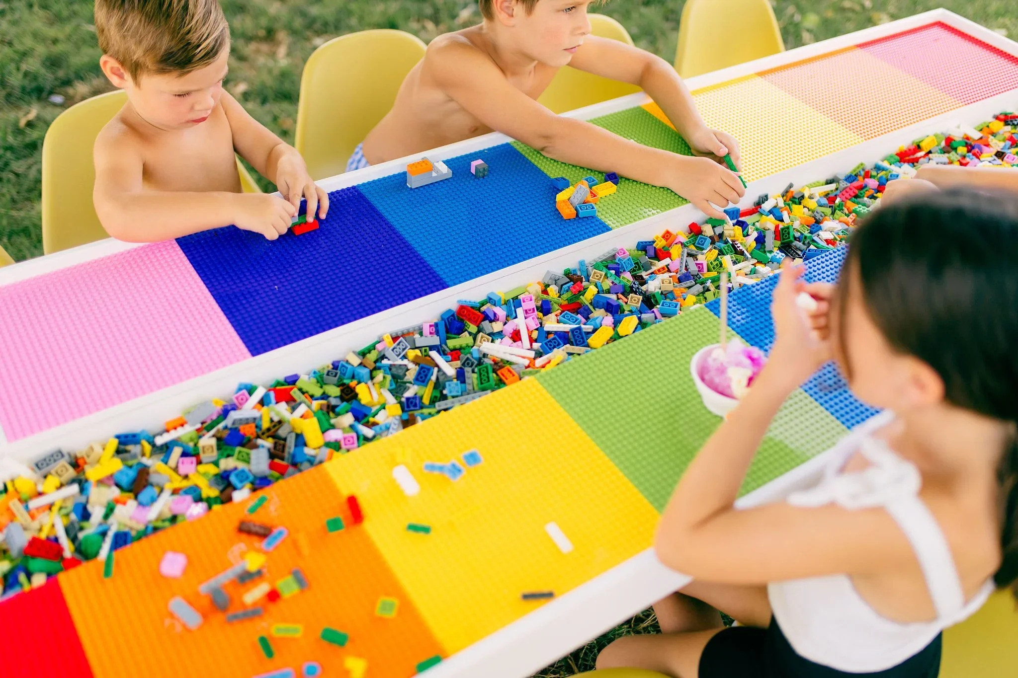 Three children sit at a long table with colorful LEGO bricks, with one girl eating ice cream. Two young boys are building with LEGO bricks, and a girl enjoys her treat while sitting among the LEGO pieces outdoors.