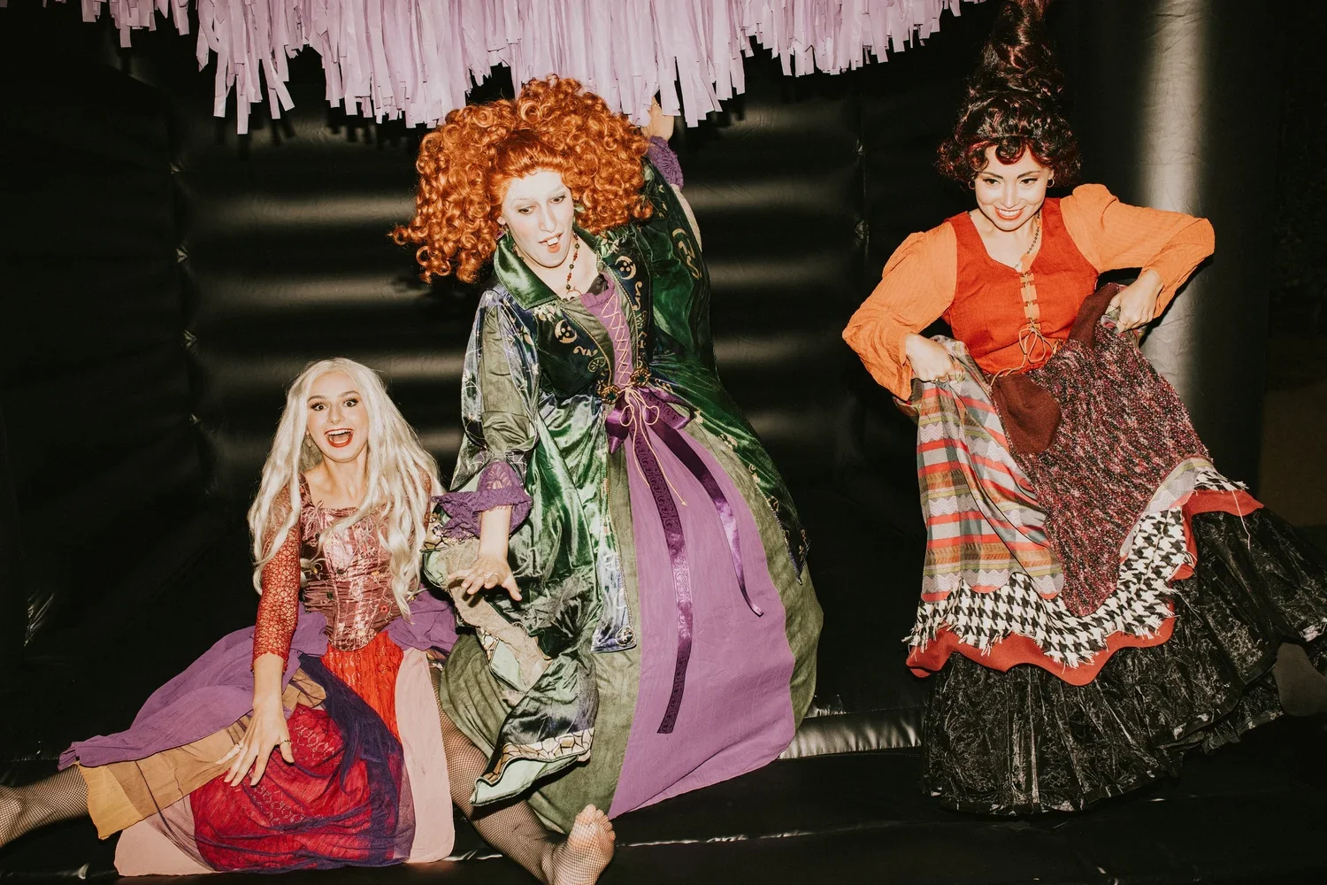 Three women in colorful, theatrical costumes sitting on a black cushioned surface with purple paper decorations hanging above them.