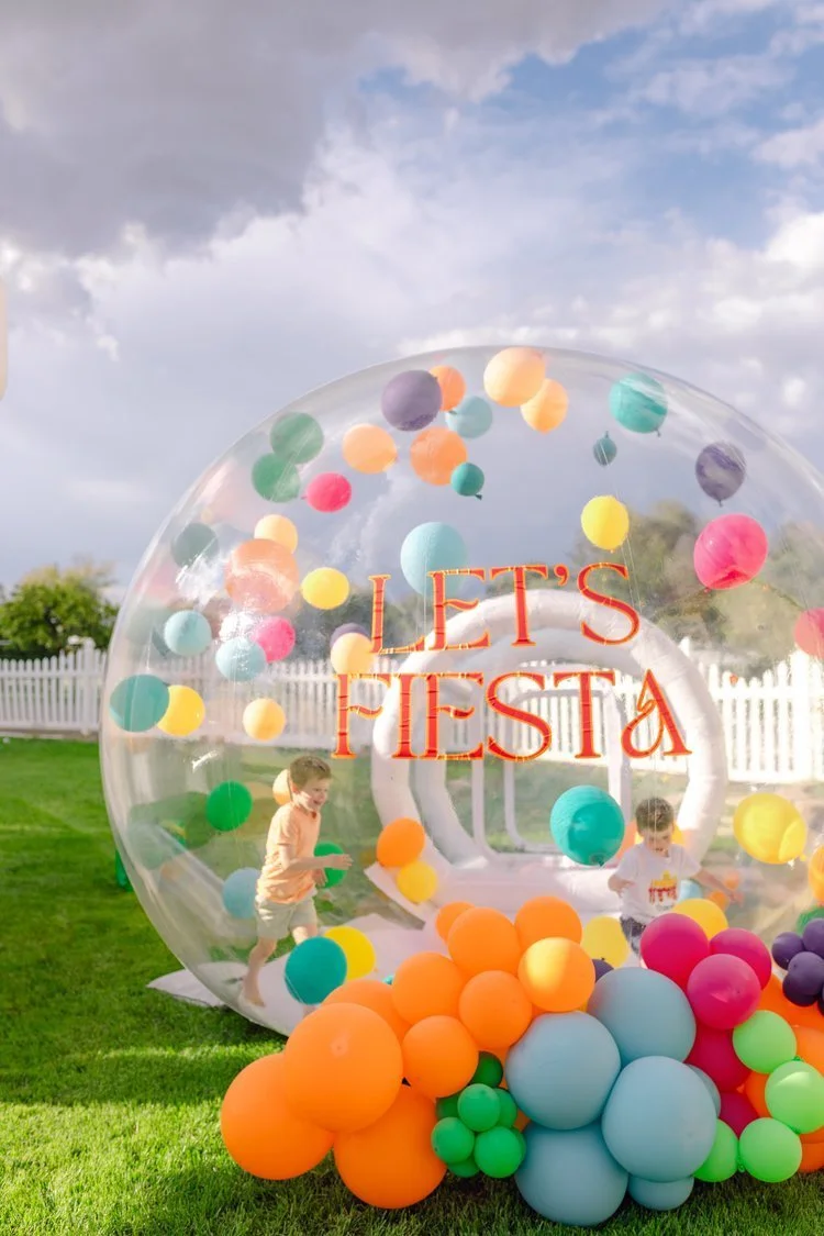 Children playing inside a clear inflatable bubble decorated with colorful balloons at a backyard party, with a white picket fence and cloudy sky in the background, and a sign reading "LET'S FIESTA" on the bubble.