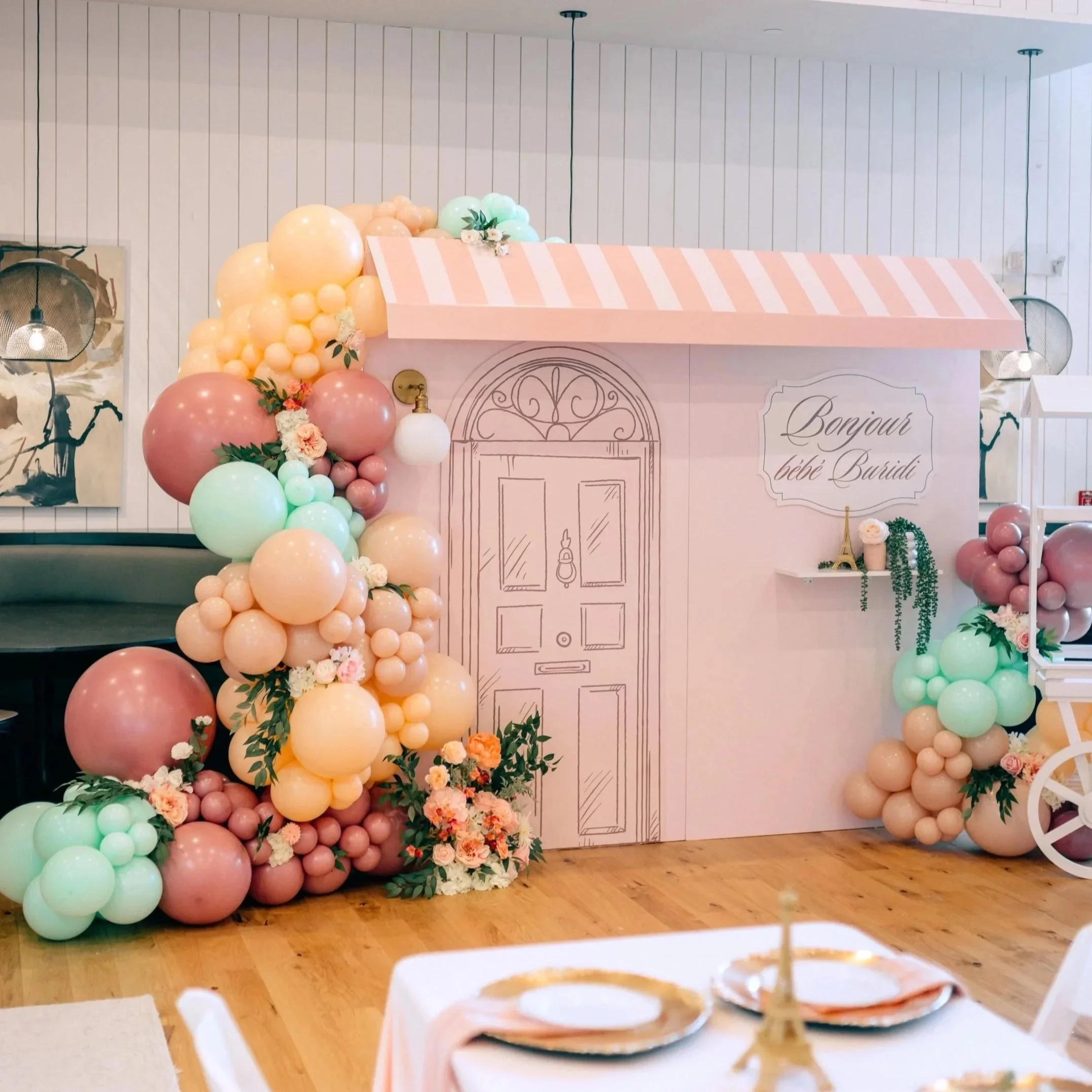 Decorative backdrop with pastel-colored balloons, flowers, and a hand-drawn door illustration for a baby shower. A sign reads 'Bonjour bébé Buridi'.