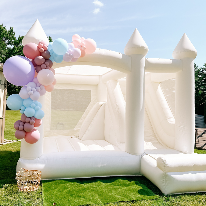 White inflatable castle with pastel balloon decorations on the left side, set up outdoors on grass under a blue sky.
