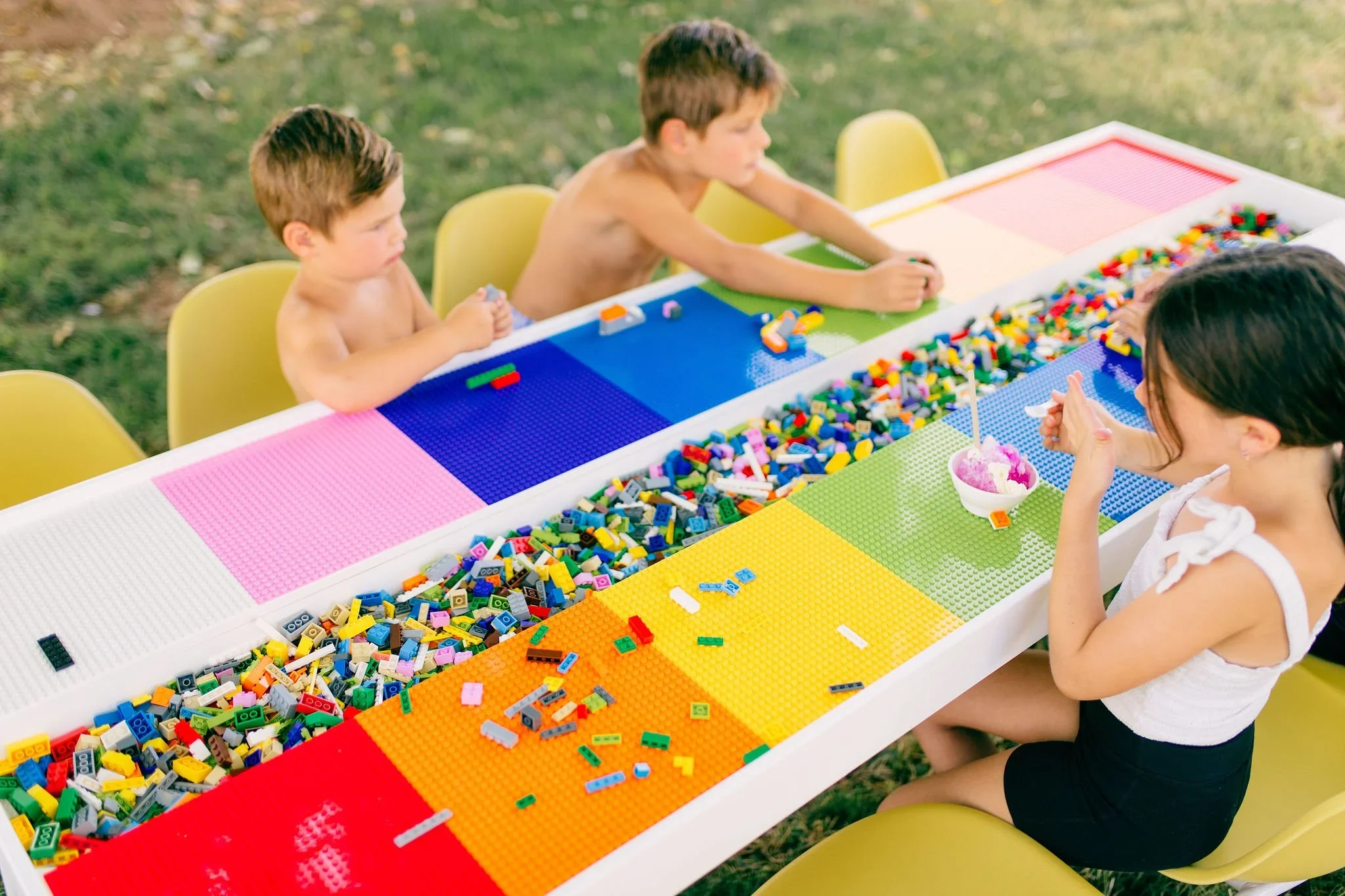 Three children and one woman sit at a long table outdoors, building with colorful LEGO bricks organized by color into sections.